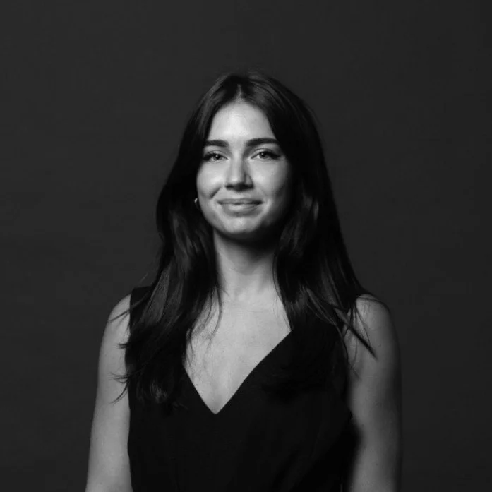 Black and white portrait of a young woman with long dark hair, smiling slightly, wearing a sleeveless black top against a dark background.