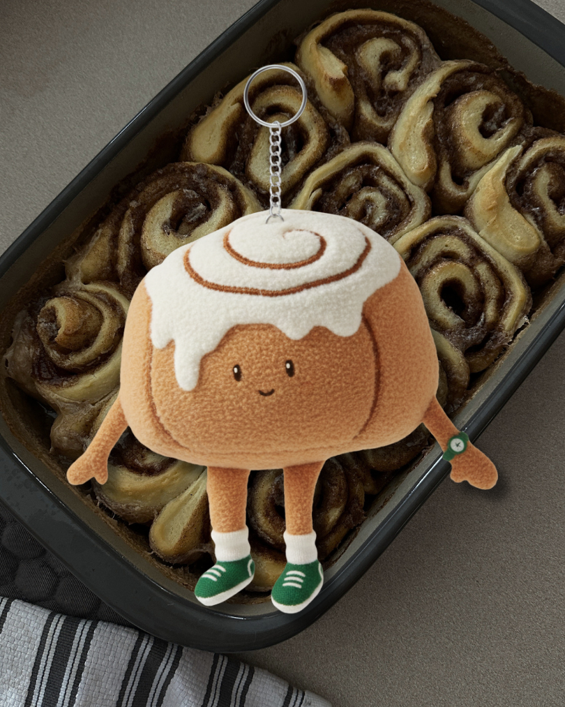 A plush cinnamon roll keychain with a cute, smiling face, green shoes, and white socks, hanging over a baking dish of cinnamon rolls.