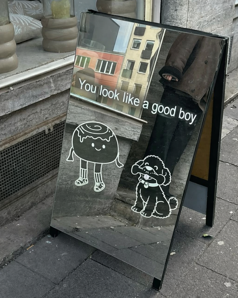 A sidewalk chalkboard sign with a reflective mirror surface, showing cartoon characters of a smiling donut with striped socks and a smiling dog with a bowtie, along with the text 'You look like a good boy' in white.