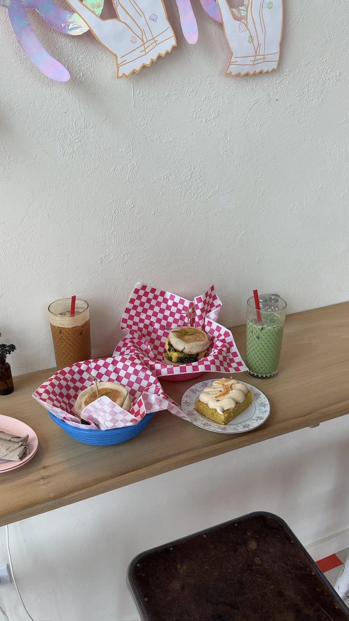 A table with various food and drinks, including sandwiches, a slice of cake, and smoothies in colorful glasses. There are pink checkered wrappers and a plate with a pastry on it. The background features a plain white wall and a wooden surface.