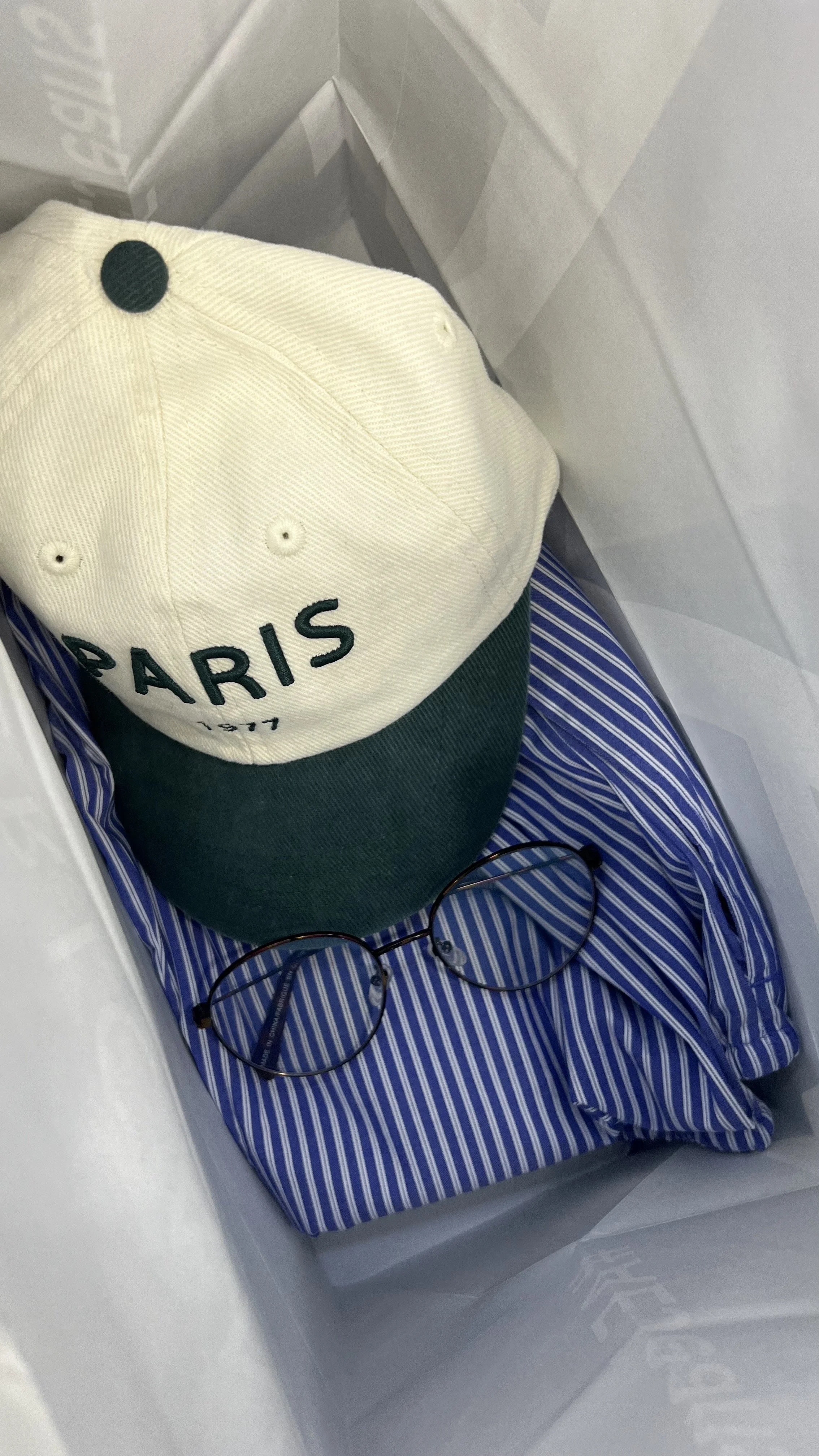 A white and green Paris baseball cap, a pair of round glasses, and a blue and white striped shirt inside a shopping bag.