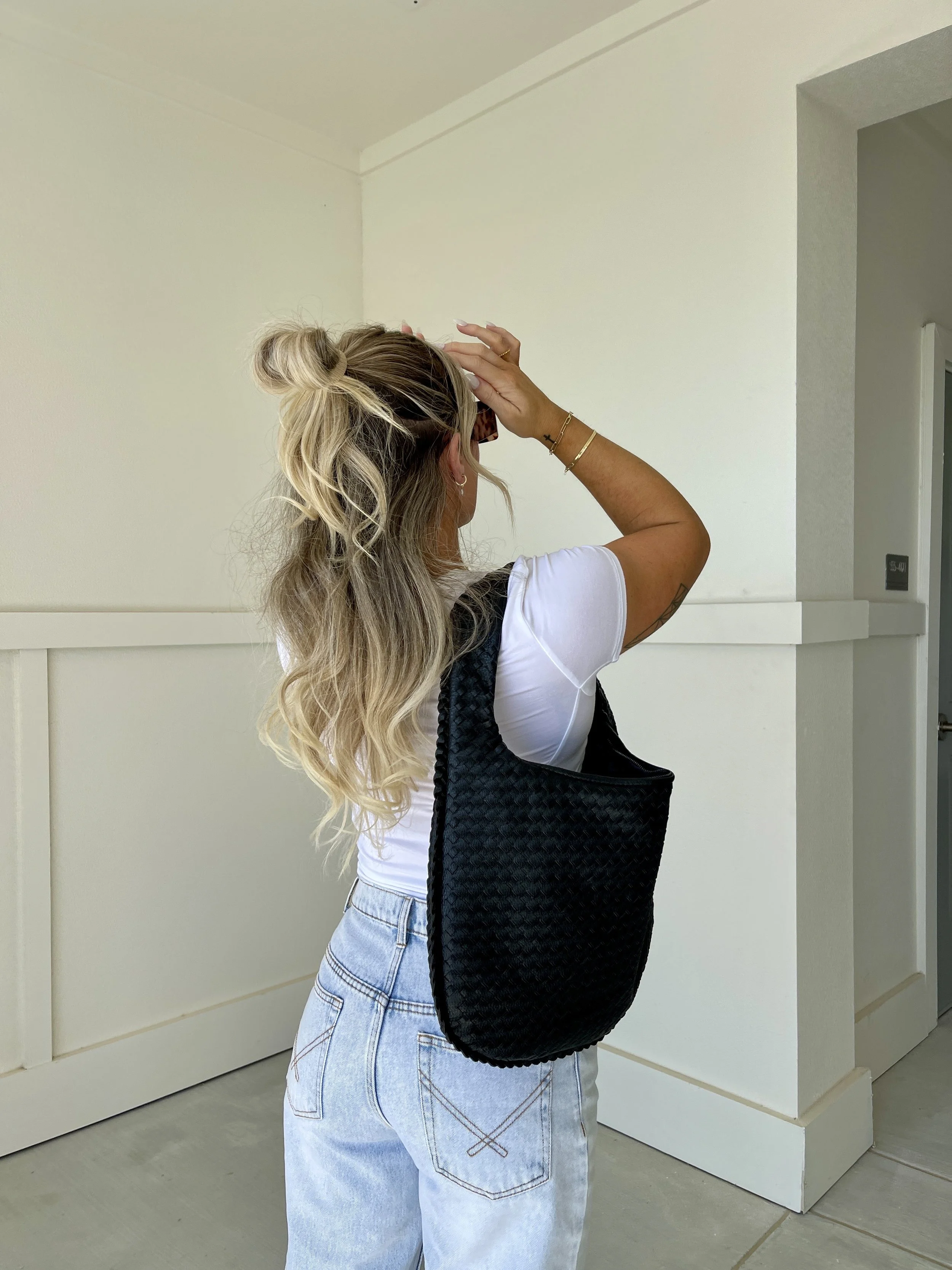Woman with blonde, wavy hair in a bun, wearing a white t-shirt and light blue jeans, carrying a black woven shoulder bag, standing indoors against a white wall.