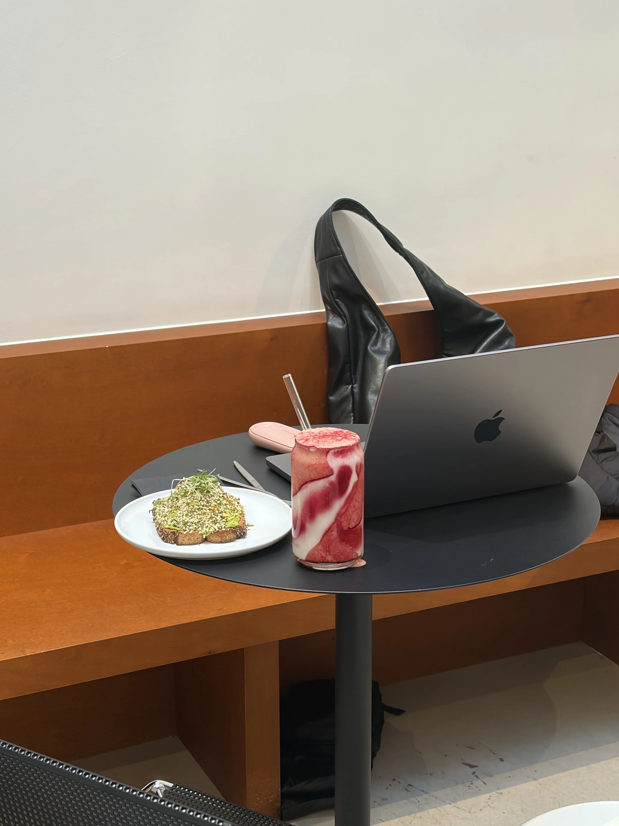 A black round table with a pink and white marble-patterned drink, a plate of food, and a silver MacBook laptop in front of a wooden booth with a black backpack hanging behind it. The background features a white wall.