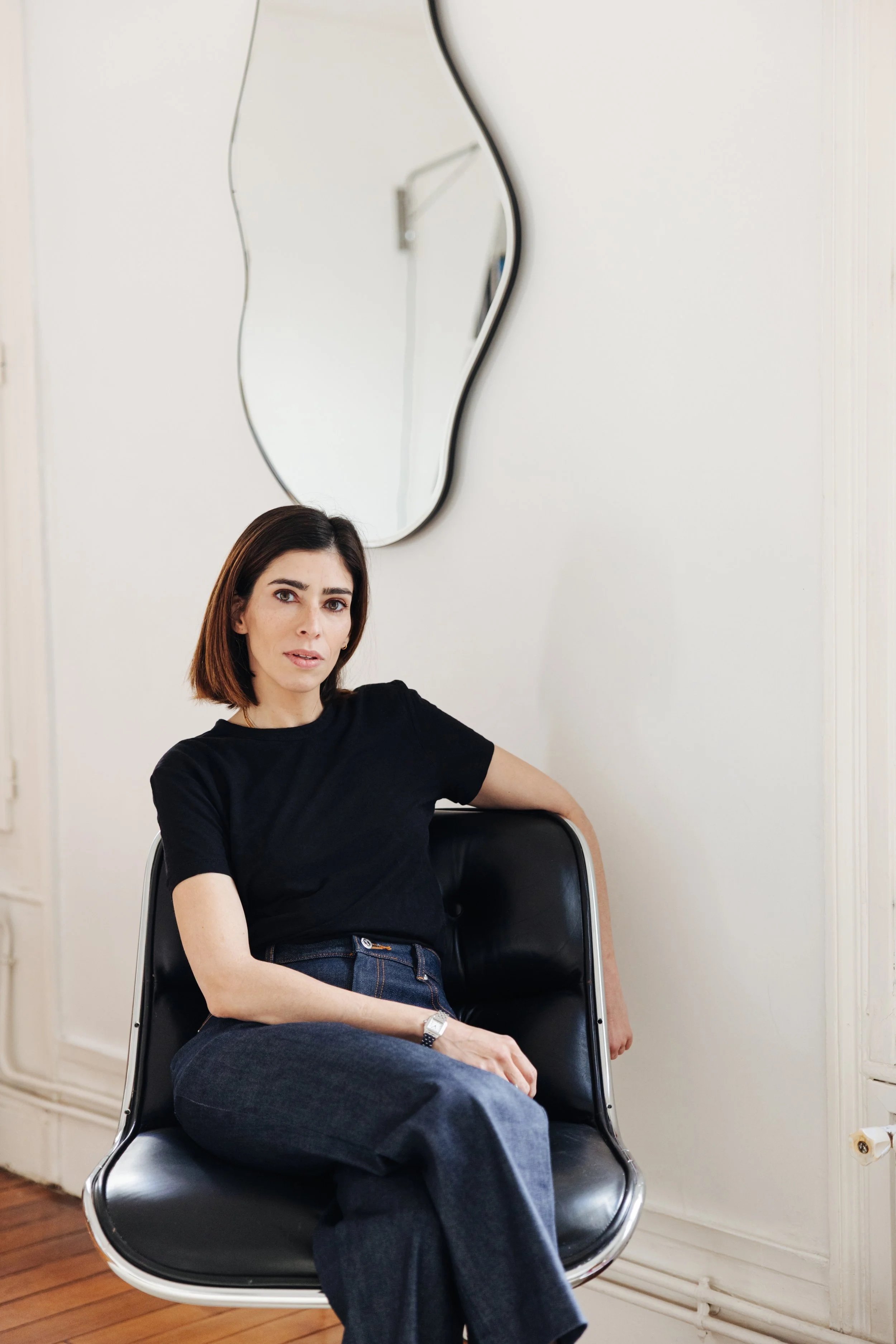 A woman with dark hair and a black t-shirt sitting on a black chair in a room with white walls and a wooden floor. There is a small mirror hanging on the wall behind her and a power outlet near the floor.