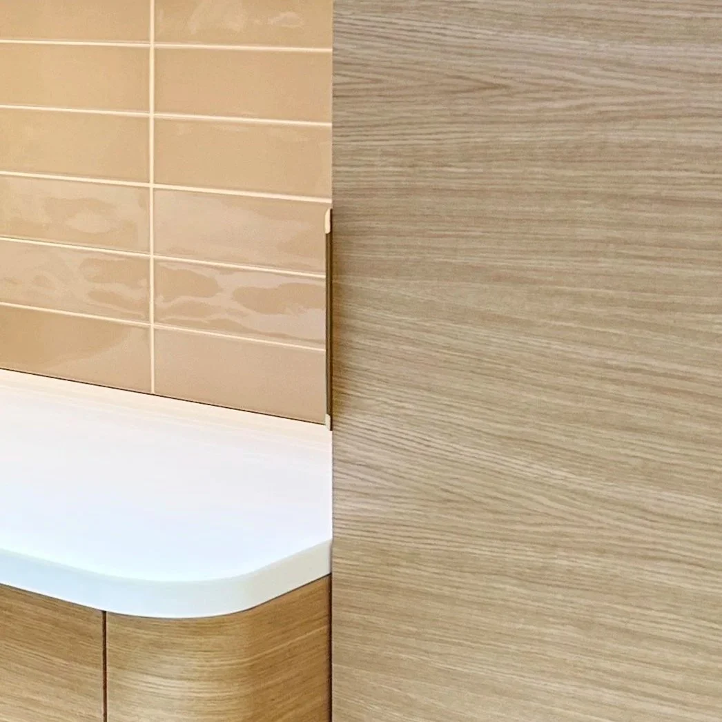 Close-up of a corner of a wooden cabinet with light wood grain, adjacent to a beige tiled wall with glossy finish and white grout lines, and a white countertop with rounded edge.