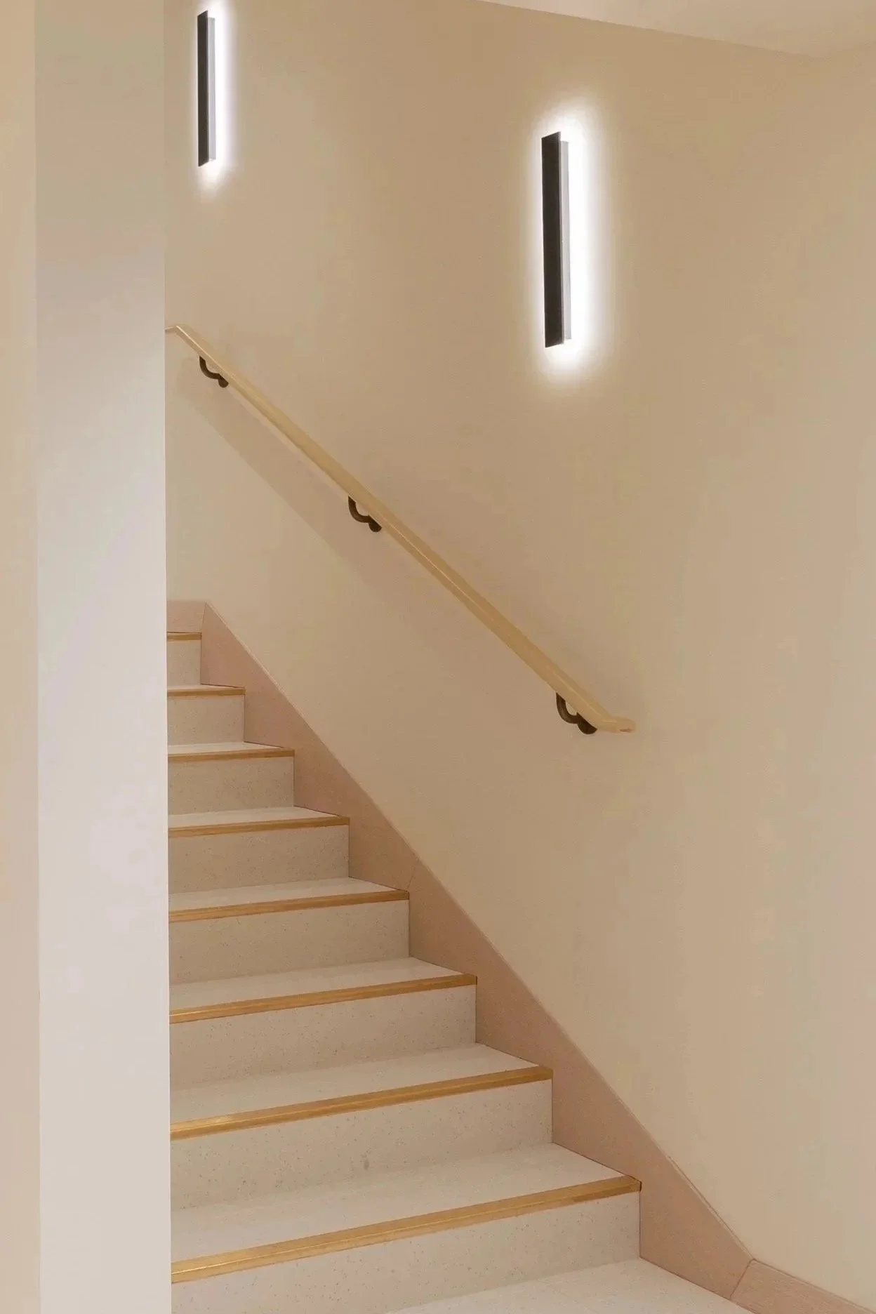 Indoor staircase with beige steps and a light-colored handrail, wall-mounted black rectangular light fixtures with backlighting, beige walls, and a beige baseboard at the bottom.