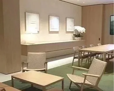 A well-lit, modern interior with a long white table and several chairs around it, a low shelf with a potted plant and framed artwork on the wall.