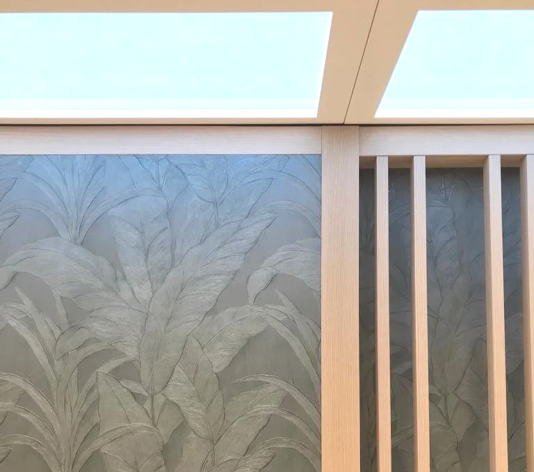 A section of an interior wall with decorative plant leaf pattern wallpaper, wooden vertical slats on the right, and a ceiling with illuminated panels.