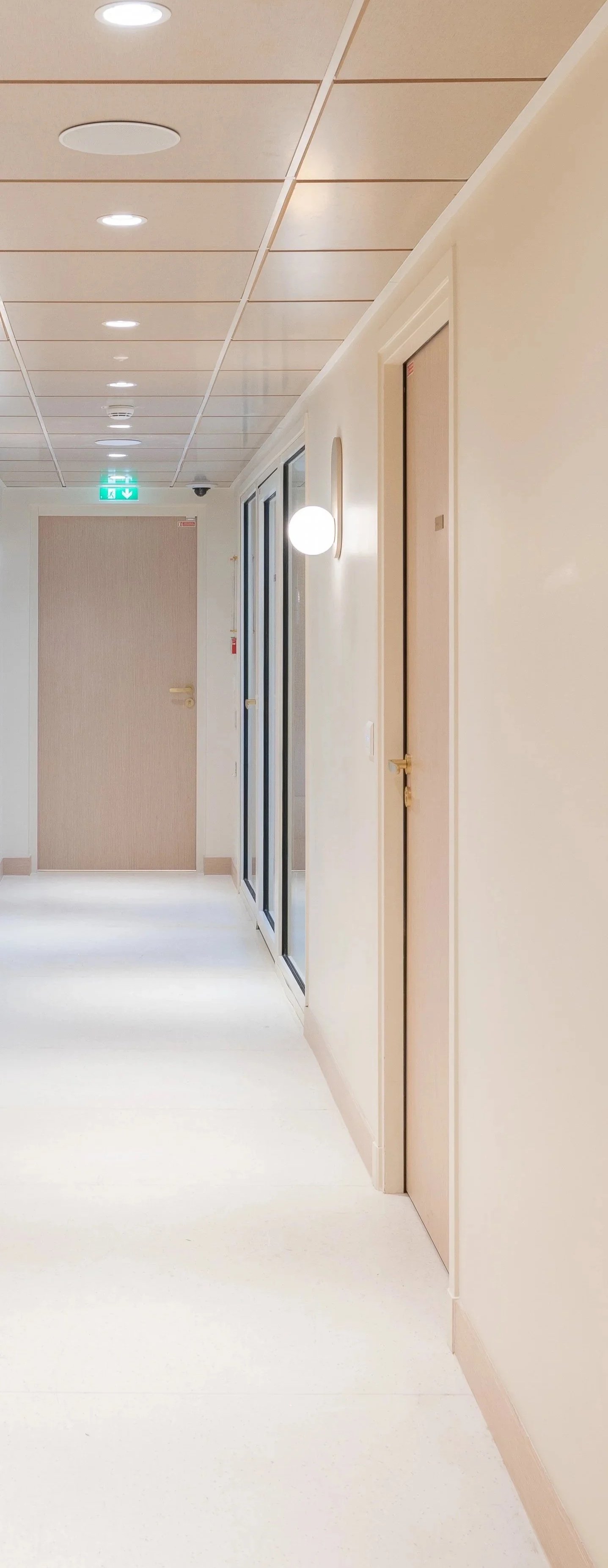 A clean hallway with beige doors, glass panels, modern white wall lights, and a bright exit sign at the end of the corridor.