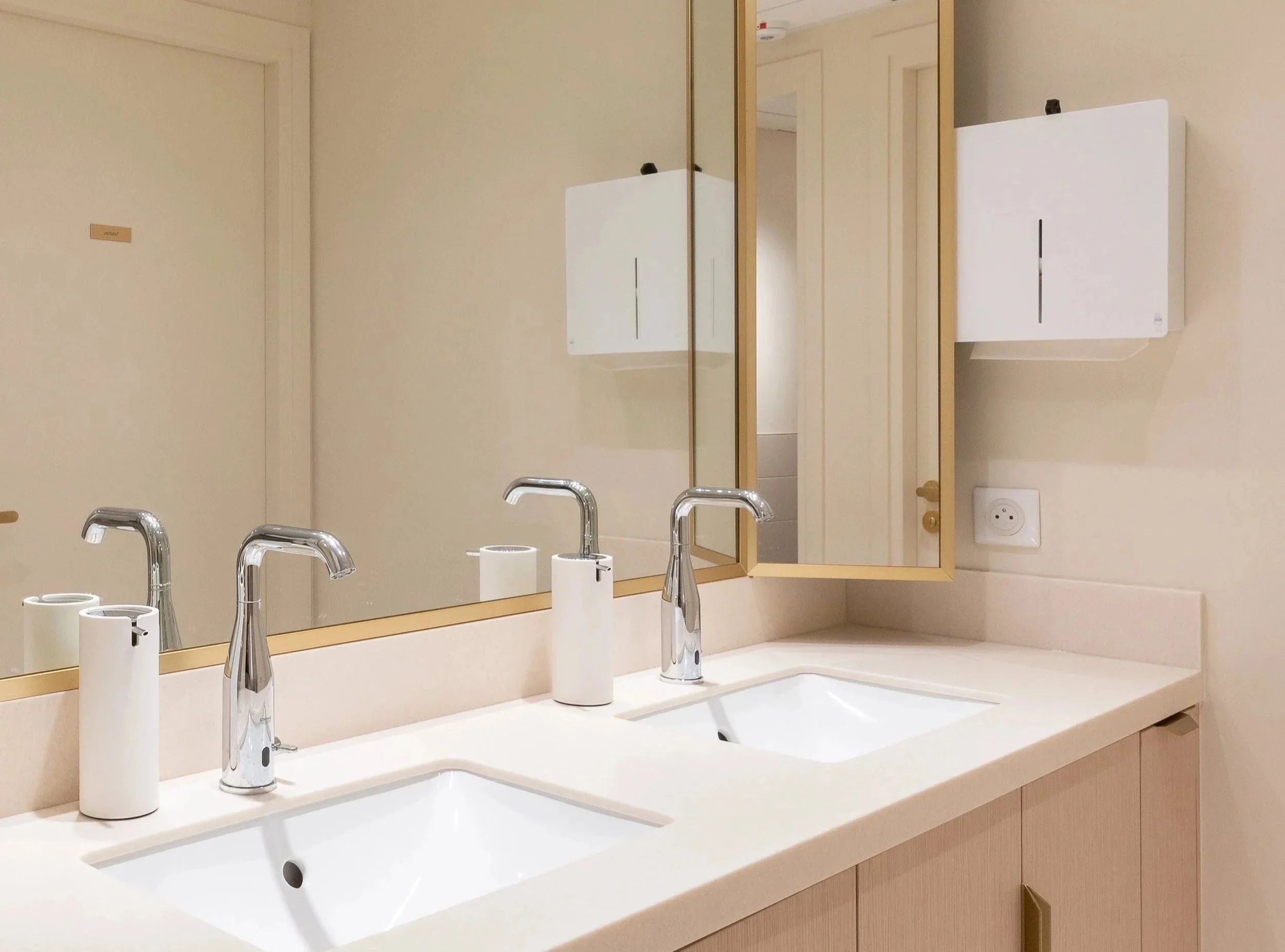 A bathroom sink with a white countertop, two silver faucets, and a large mirror above it. The mirror has a gold-colored frame and a smaller tilting mirror attached to it. The wall behind is beige, and there are two white paper towel rolls and a soap 