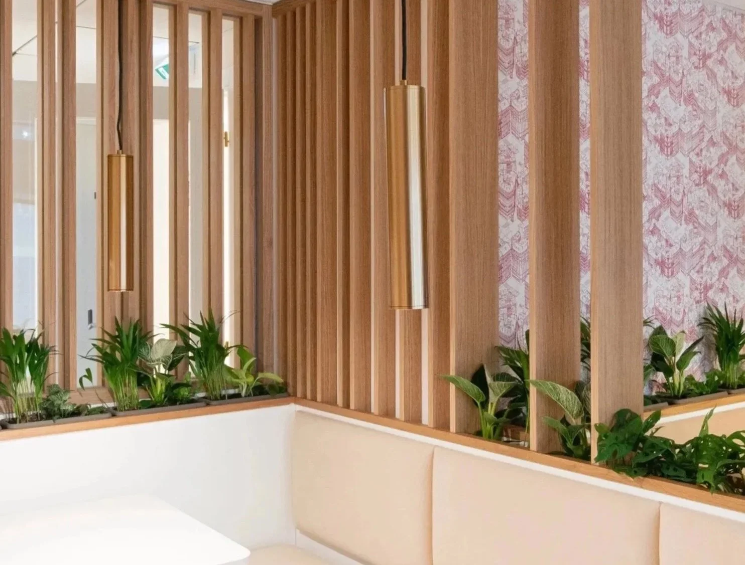 Interior scene with vertical wooden slats partition, pink patterned wallpaper on the wall, hanging gold pendant lights, and green potted plants along the ledge.