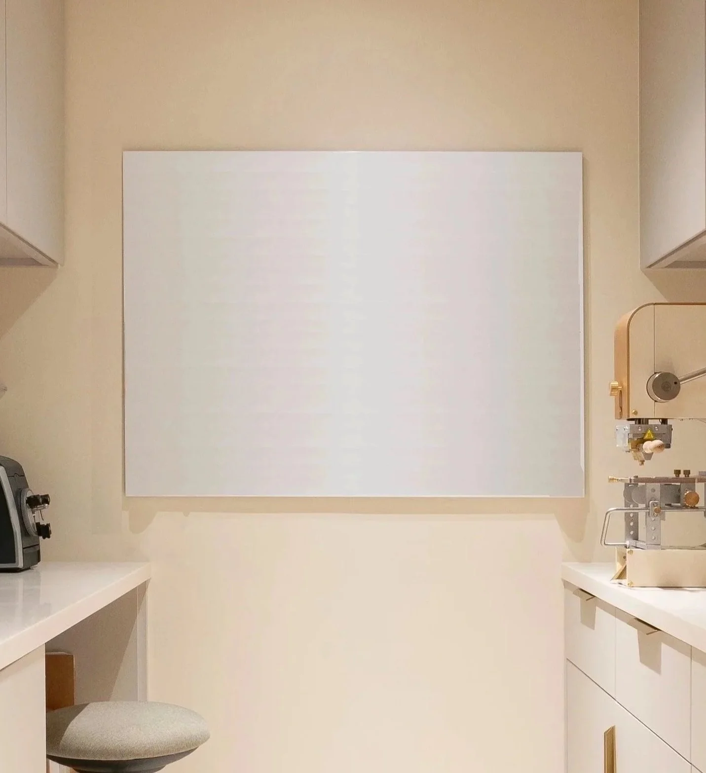 A small, minimalist medical or scientific laboratory room with a white counter, a gray stool, built-in cabinets with gold handles, and laboratory equipment attached to the wall, including a microscope and some glassware.