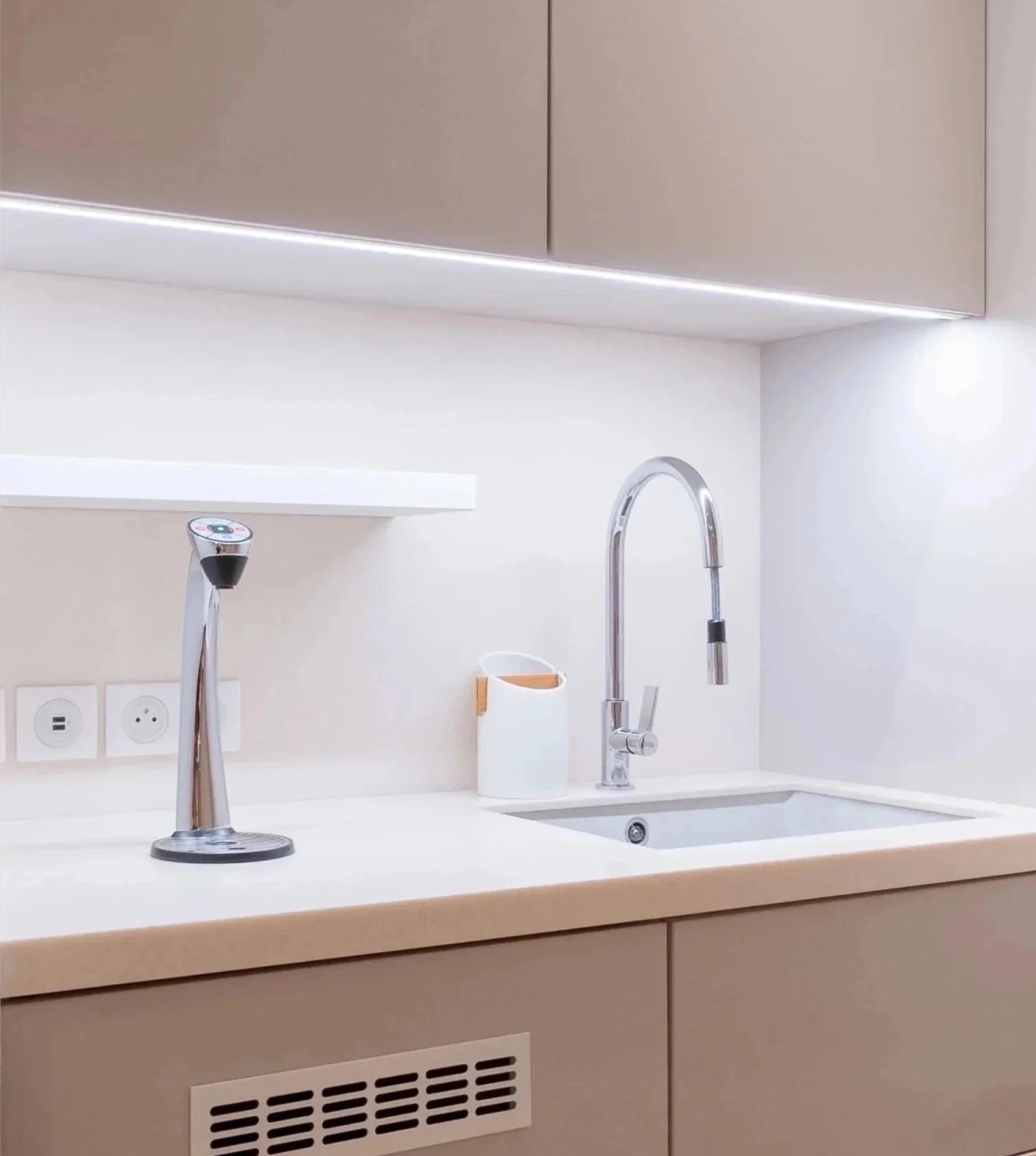 A modern kitchen with a beige cabinet, a white countertop, a sink with a high-arc faucet, and wall outlets. A soap dispenser is on the counter, and there is an under-cabinet light giving a soft glow.