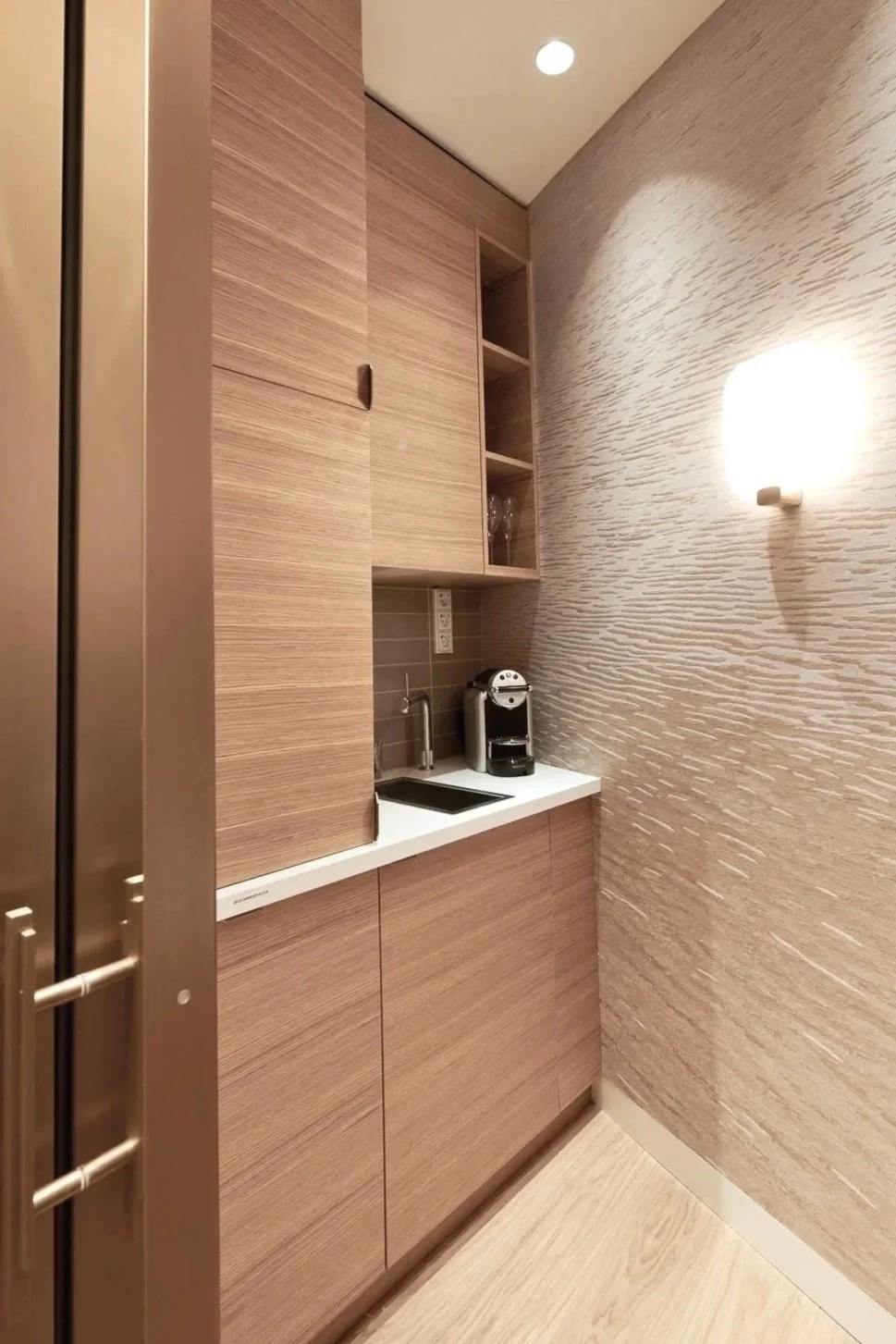 A small kitchen with beige wood cabinets, a white countertop, a coffee machine, a sink, and a wall-mounted light fixture.