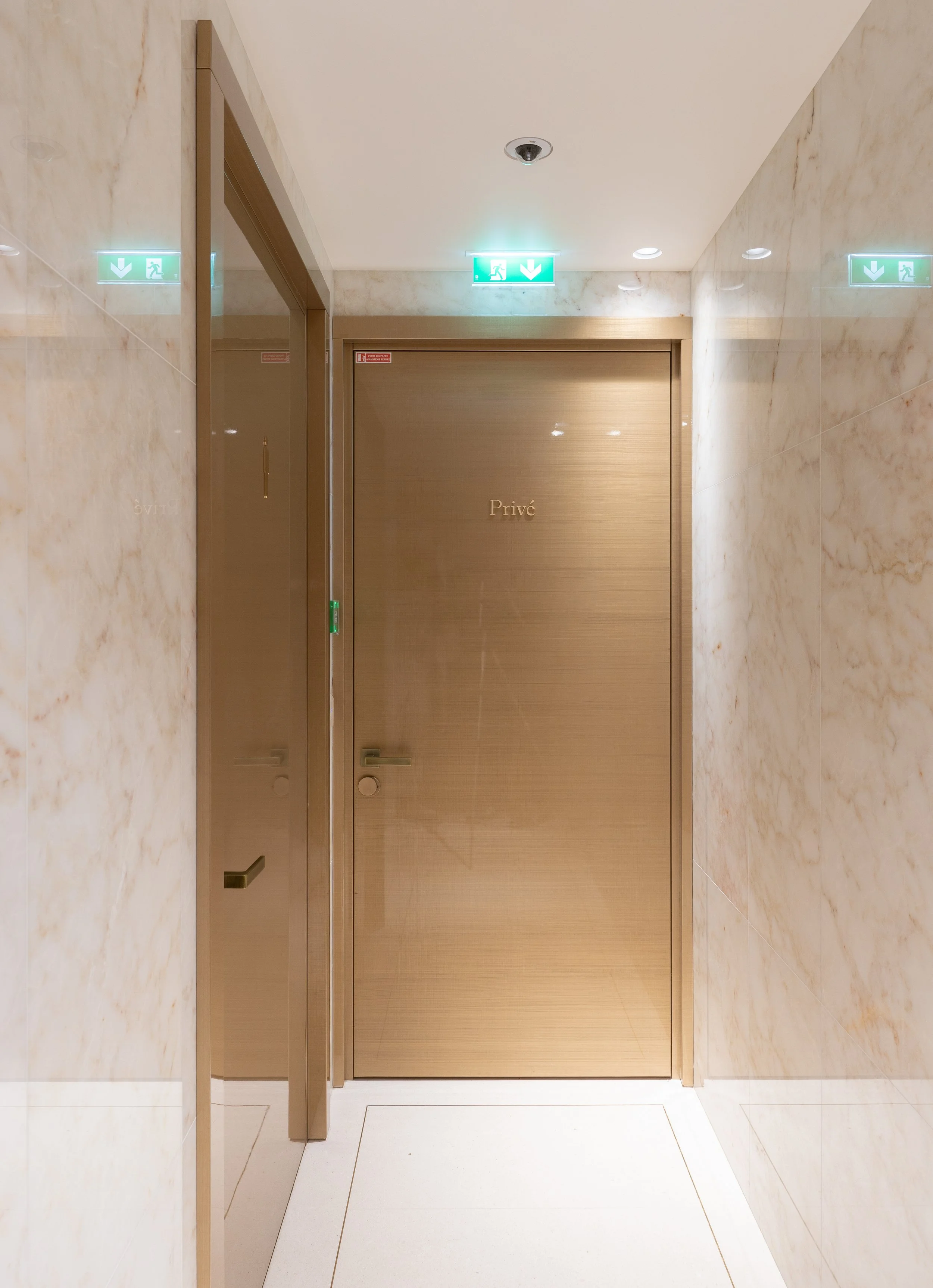 Gold-colored doors with the word 'Privé' on them, in a hallway with marble walls and white tile floor, overhead green emergency exit signs.