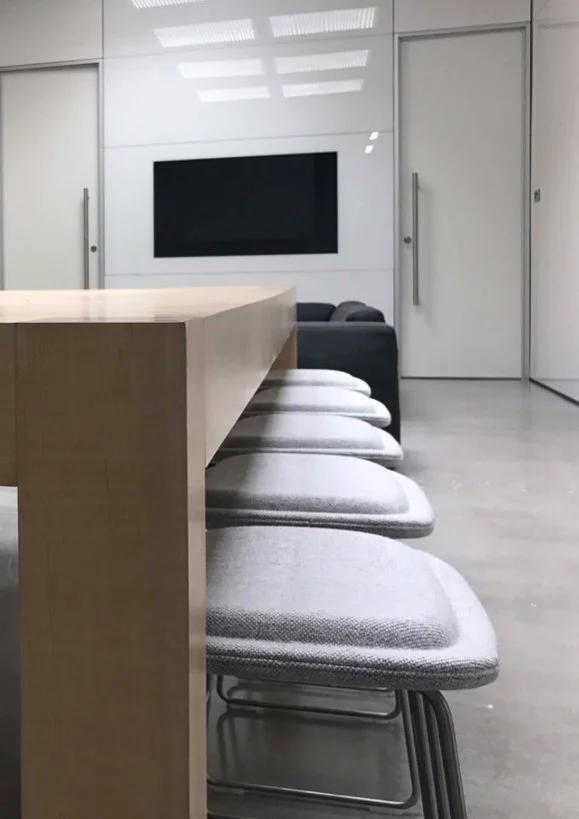Modern conference room with a wooden table, gray upholstered chairs, a black wall-mounted TV, and white walls with sliding doors.