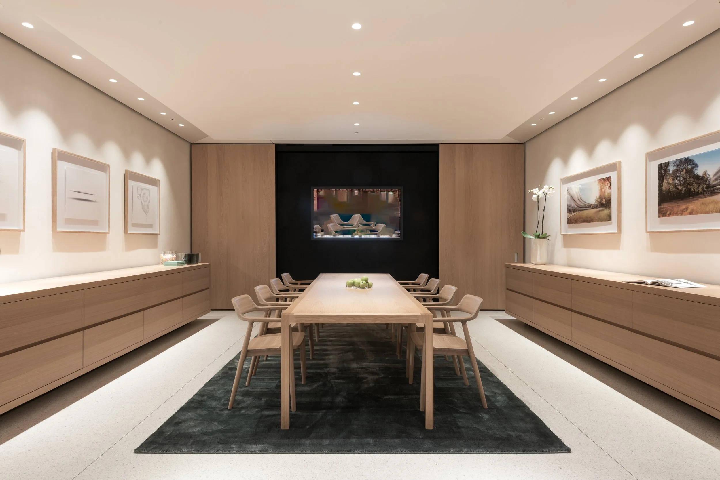A modern dining room with a long wooden table, surrounded by matching wooden chairs, with built-in wooden cabinets along both side walls. A black and white area rug runs underneath the table, and a small decorative bowl of green apples is at the cent