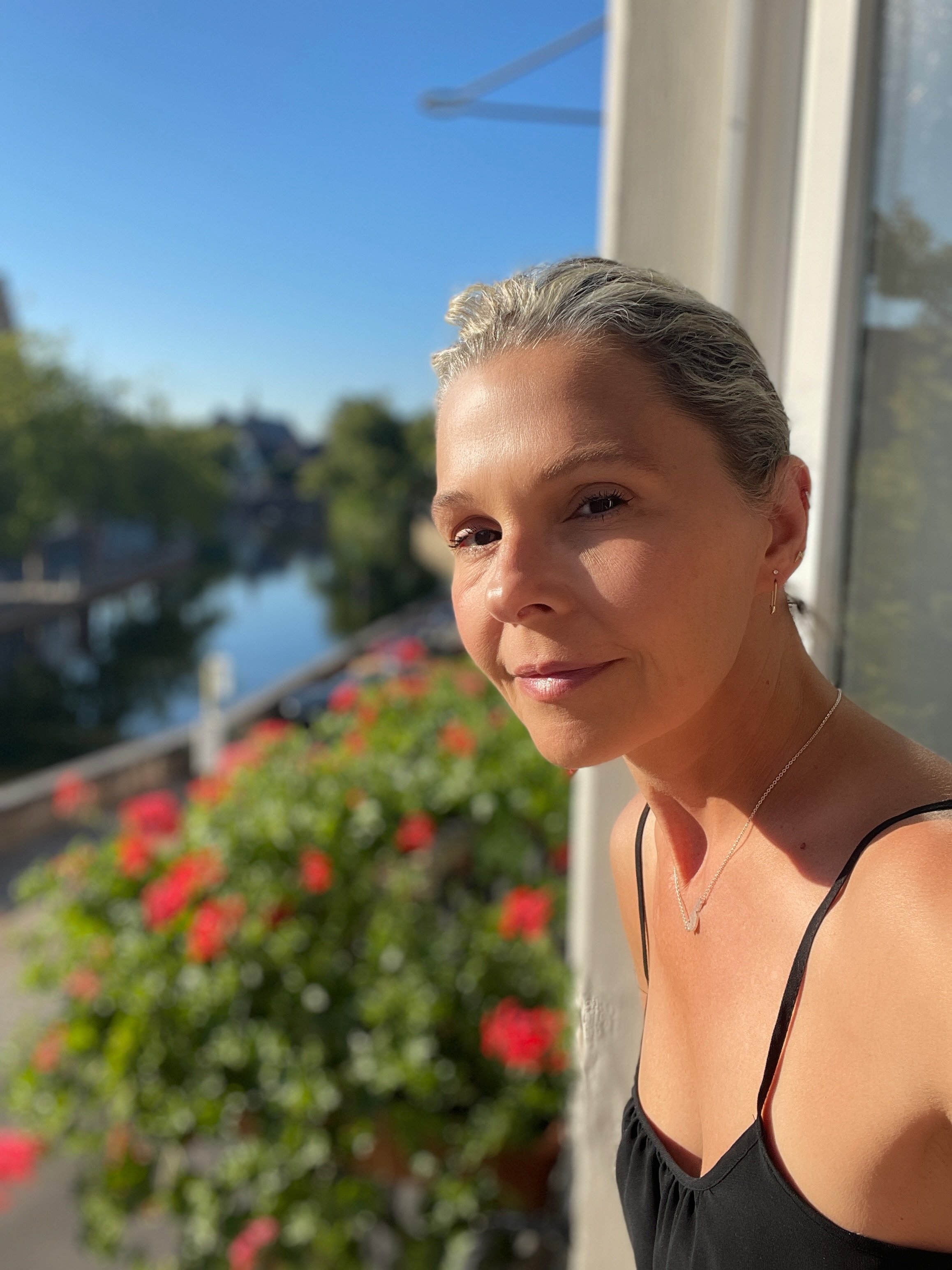 Woman with gray hair smiling outdoors on a sunny day, with a canal, trees, and colorful flowers in the background.