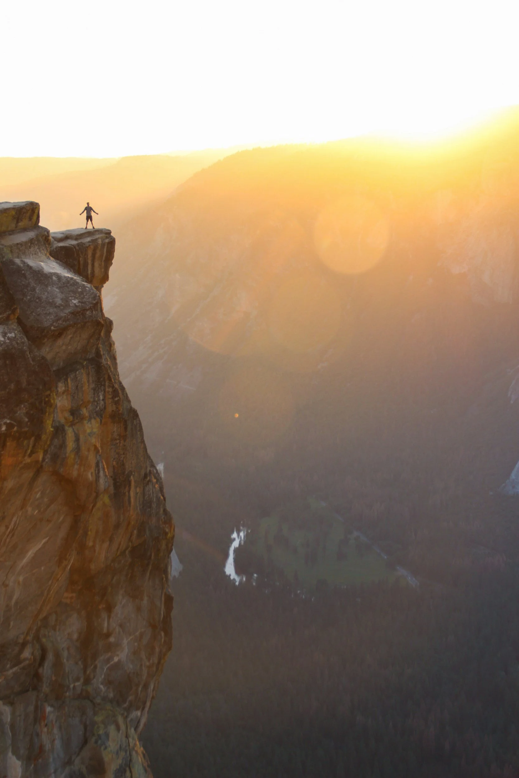 Person standing on edge of a high cliff at sunrise, overlooking a valley.