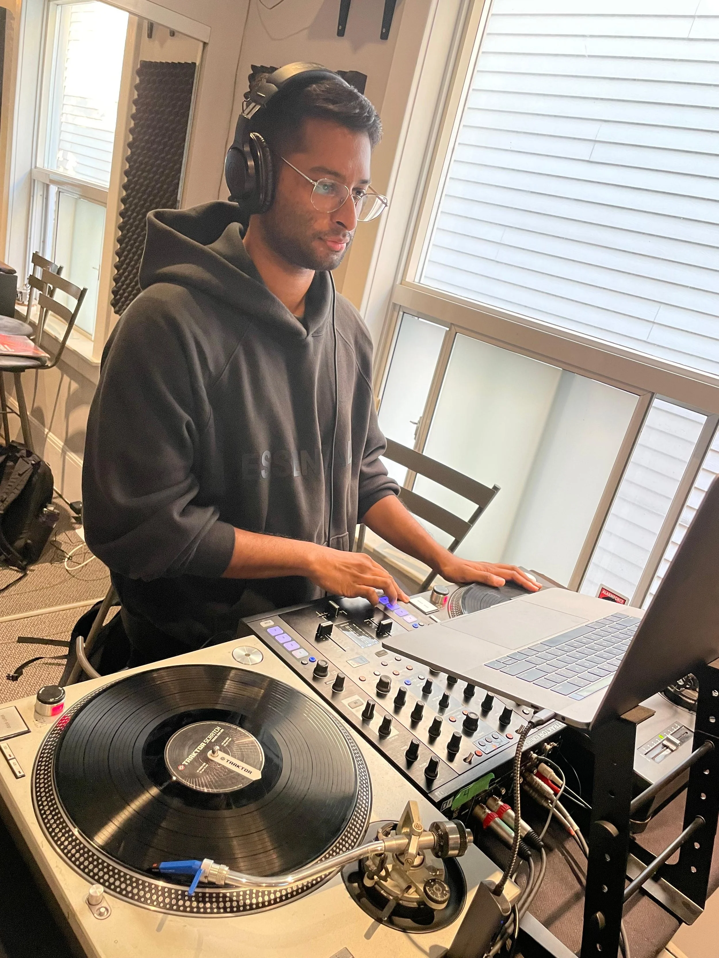 A man wearing glasses and headphones DJing with turntables, a mixer, and a laptop in a room with large windows.