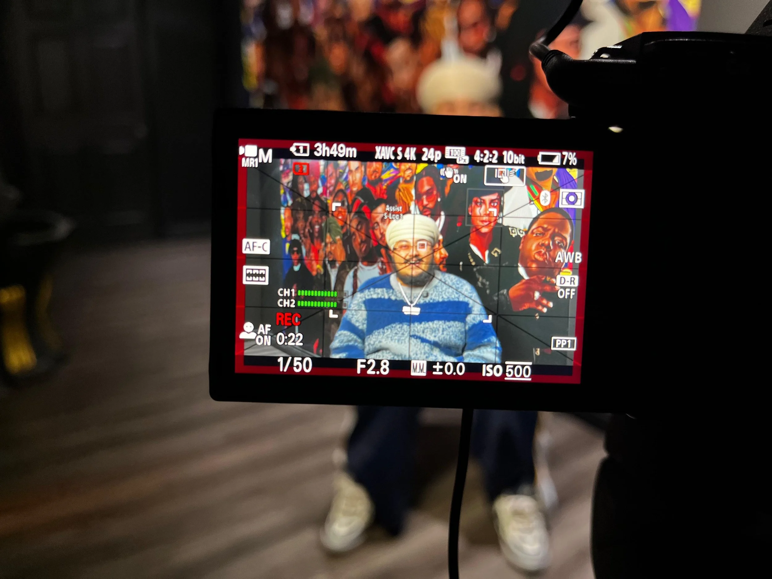 Camera screen capturing a picture of a group of people in colorful clothing, with two men in the foreground, one wearing a turban and the other with dark skin, both smiling.