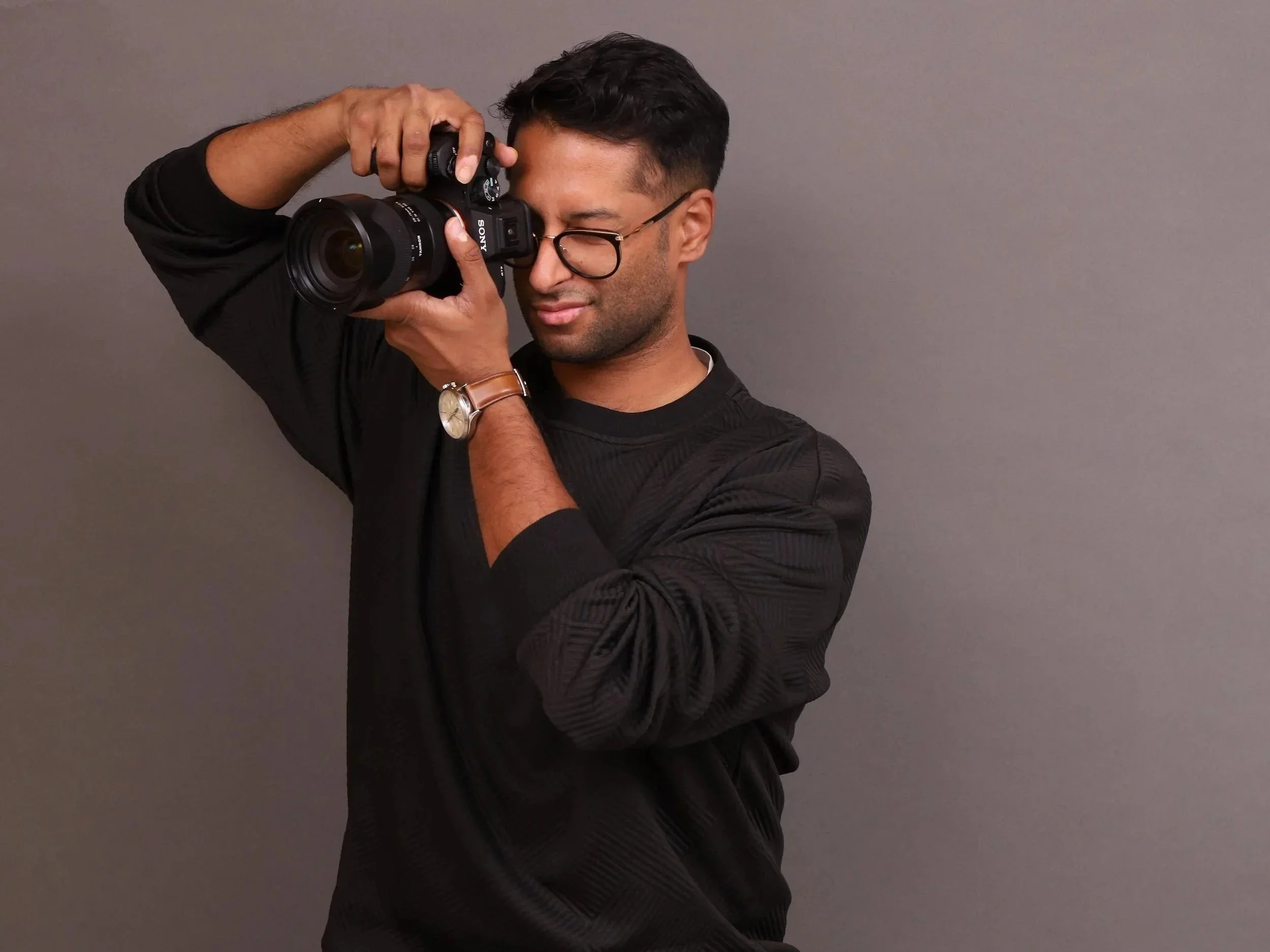 A man with dark hair, glasses, and a watch is holding a camera up to his eye, aiming it to take a photograph against a plain gray background.