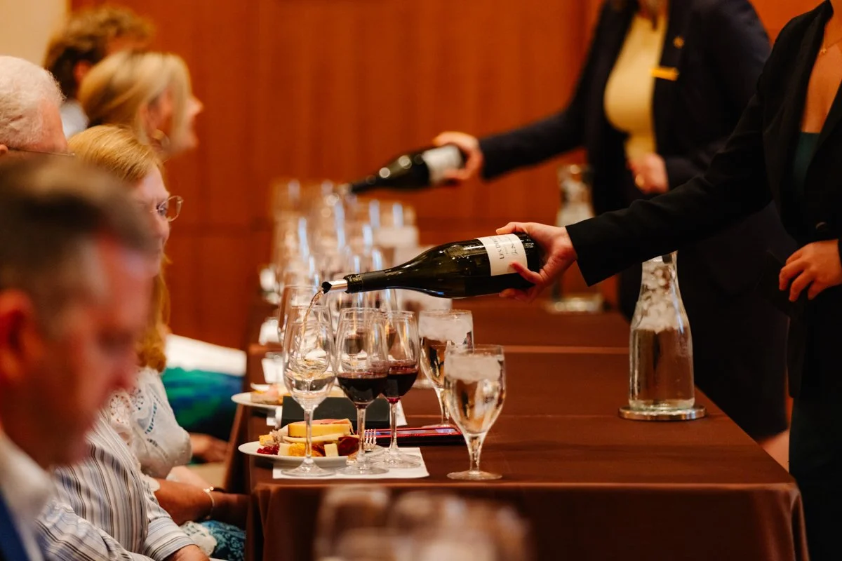 People seated at a dining table with glasses of wine and water, as waitresses pour wine during a formal event or wine tasting.