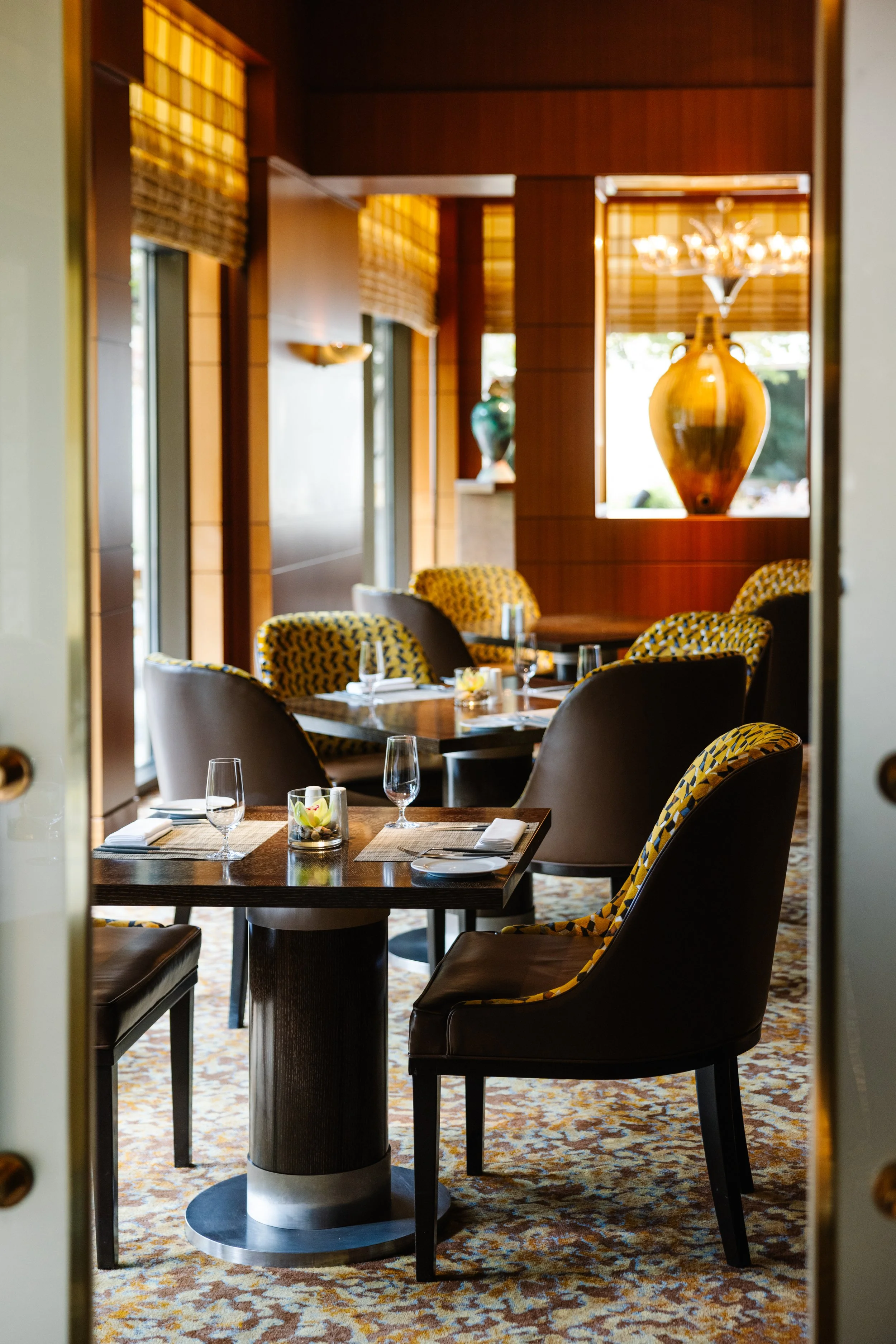 View of an elegantly decorated restaurant dining area with wooden walls, large windows with window treatments, and a chandelier. The tables are set with glasses, napkins, and small floral centerpieces. The chairs have a yellow and black geometric pattern and leather upholstery, and the room features decorative vases displayed on pedestals.