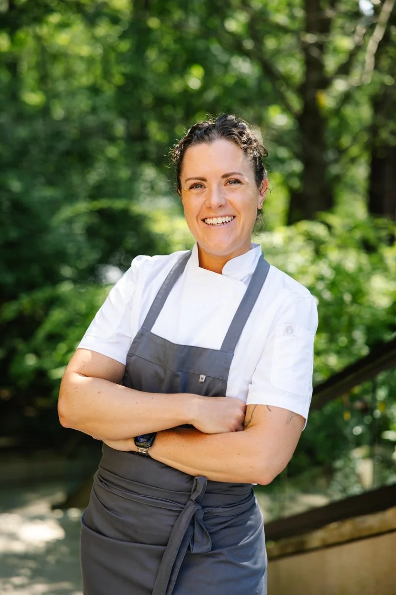 A smiling woman in a chef's coat and gray apron standing outdoors in a lush green park.