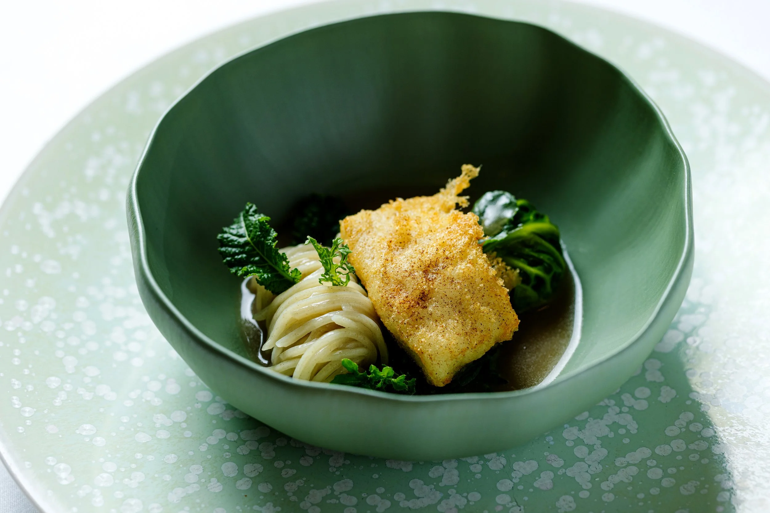 Breaded fish fillet with noodles, greens, and herbs in a green bowl.