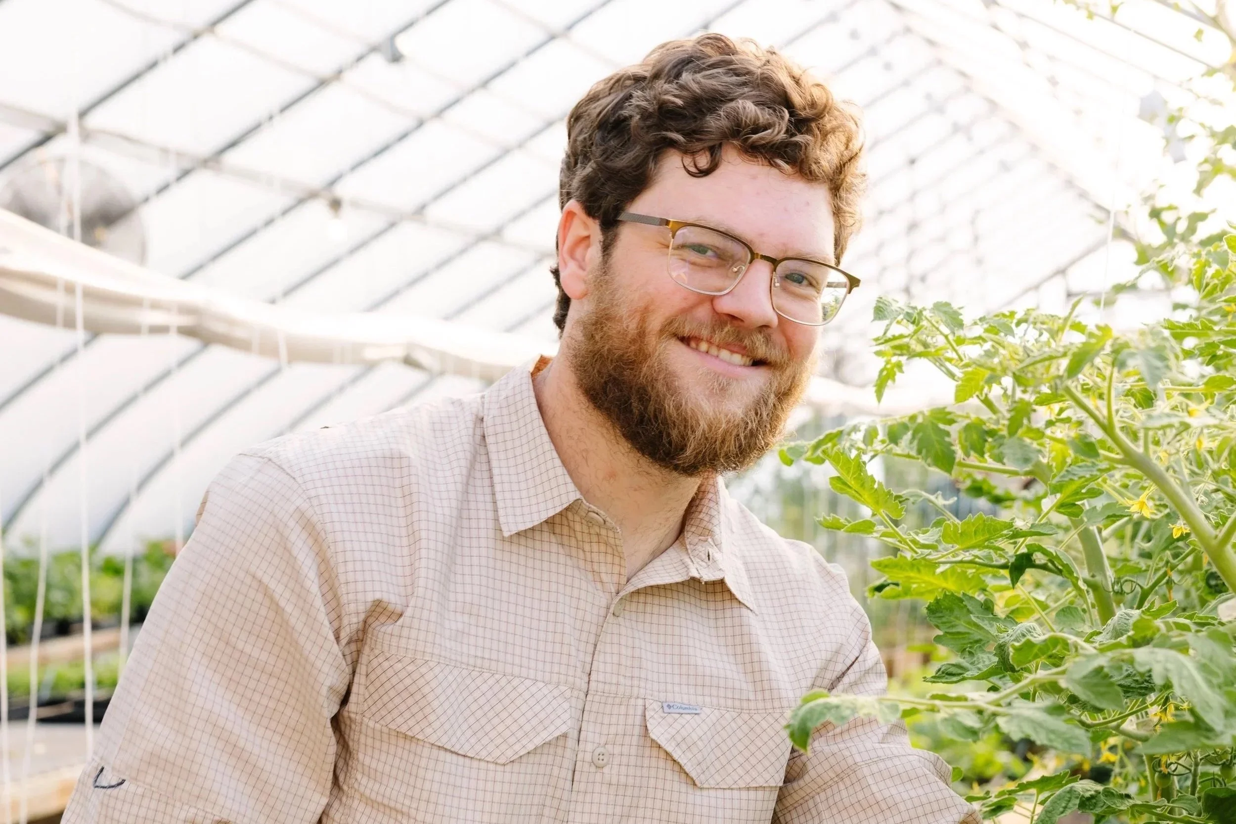 A smiling man with glasses and a beard working in a greenhouse surrounded by green plants.