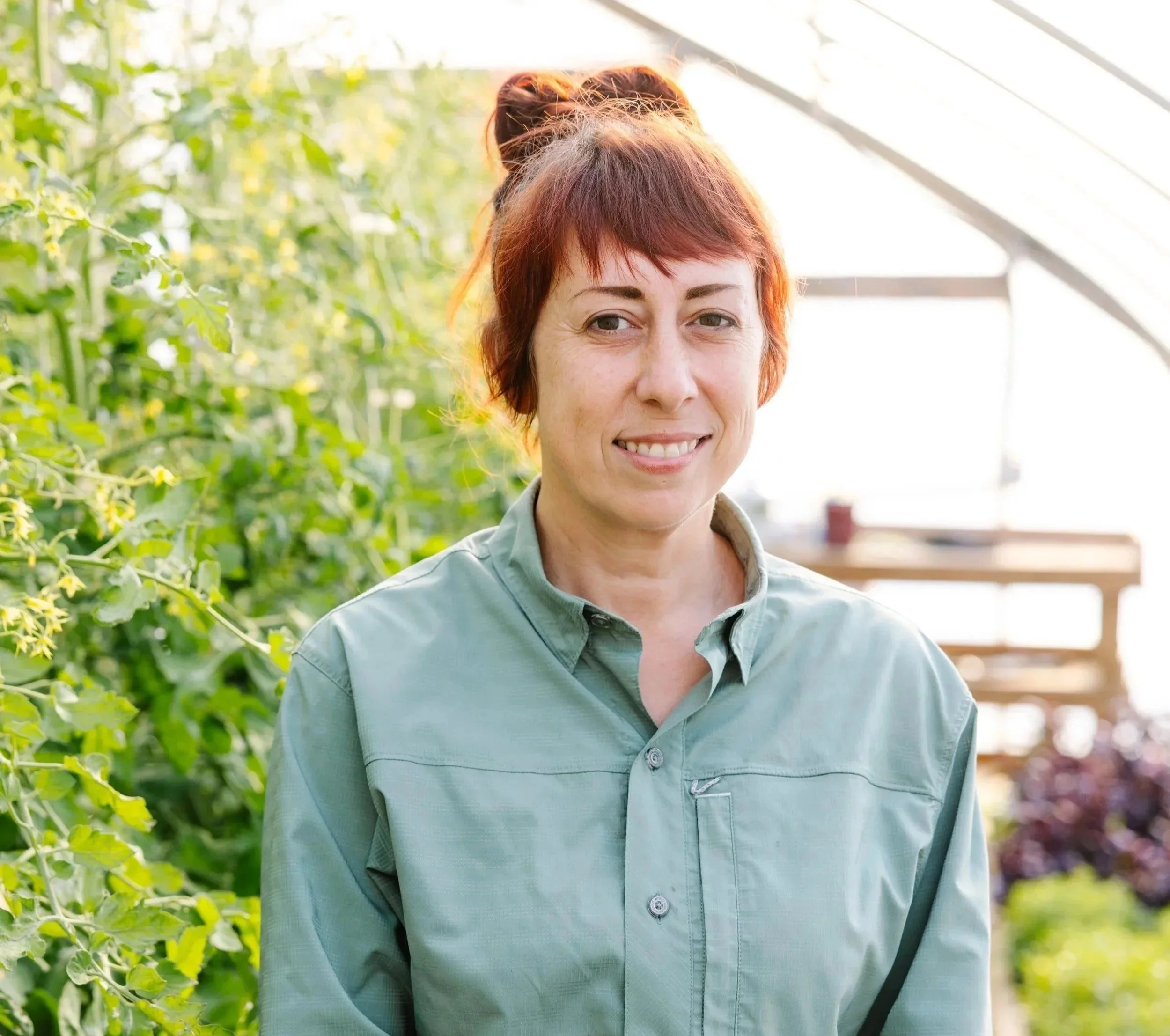 A woman with short red hair wearing a green button-up shirt standing in a greenhouse with lush green plants in the background.