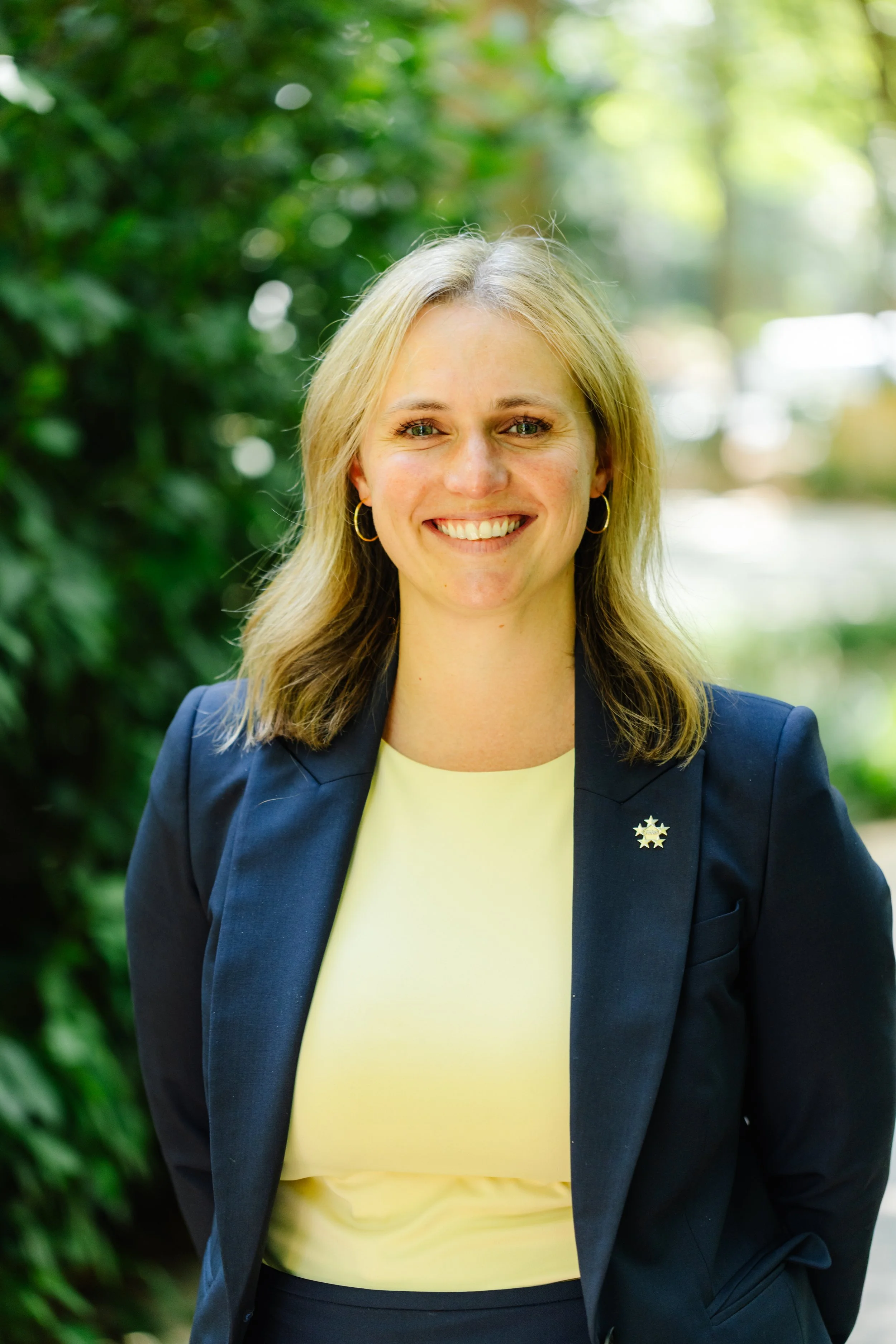 A young woman with shoulder-length blonde hair smiling outdoors, wearing a navy blazer with a star-shaped pin, a light yellow top, and small hoop earrings, with green foliage in the background.