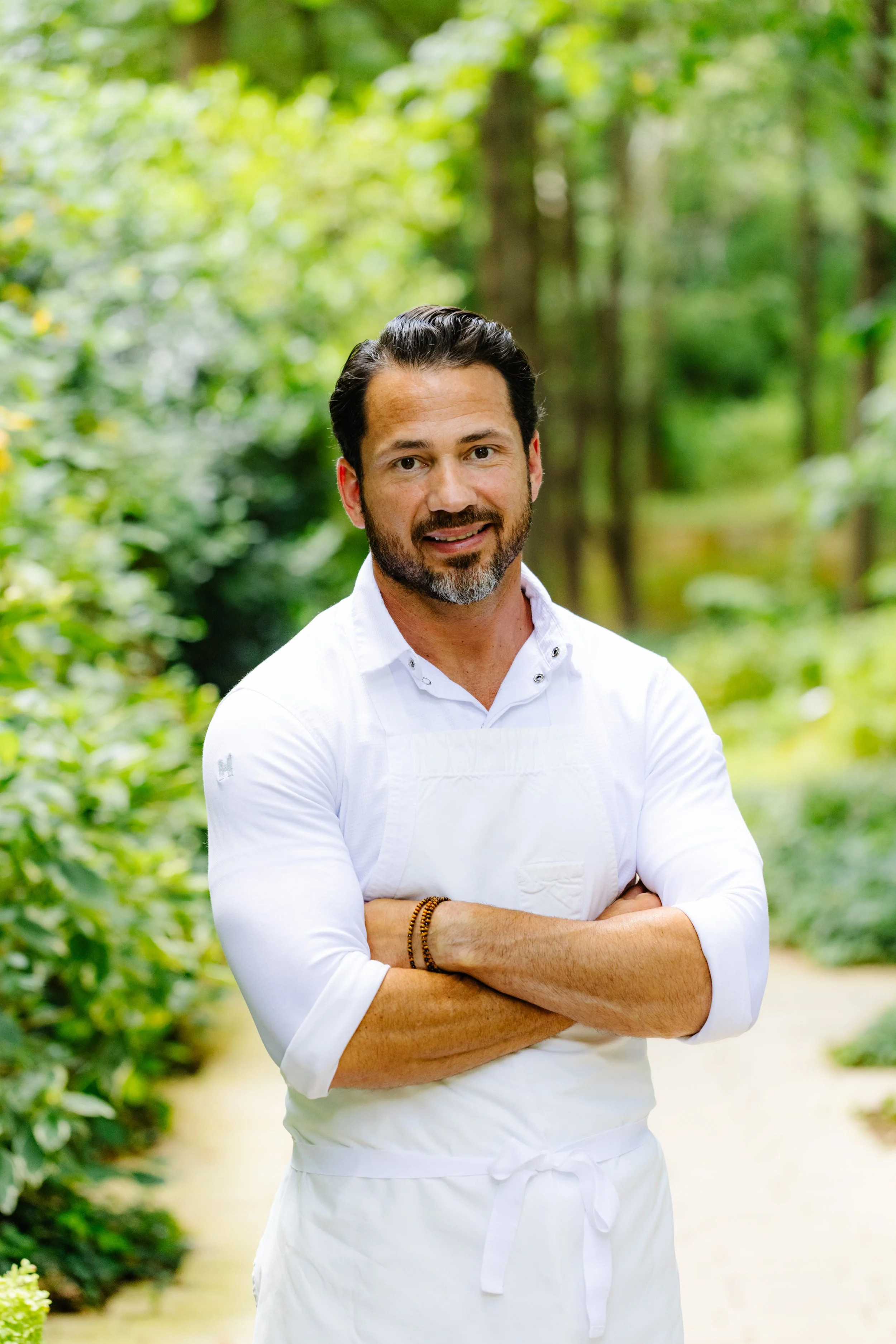 A man with dark hair and a beard, wearing a white shirt and apron, stands outdoors in a lush green forested area with his arms crossed, smiling at the camera.