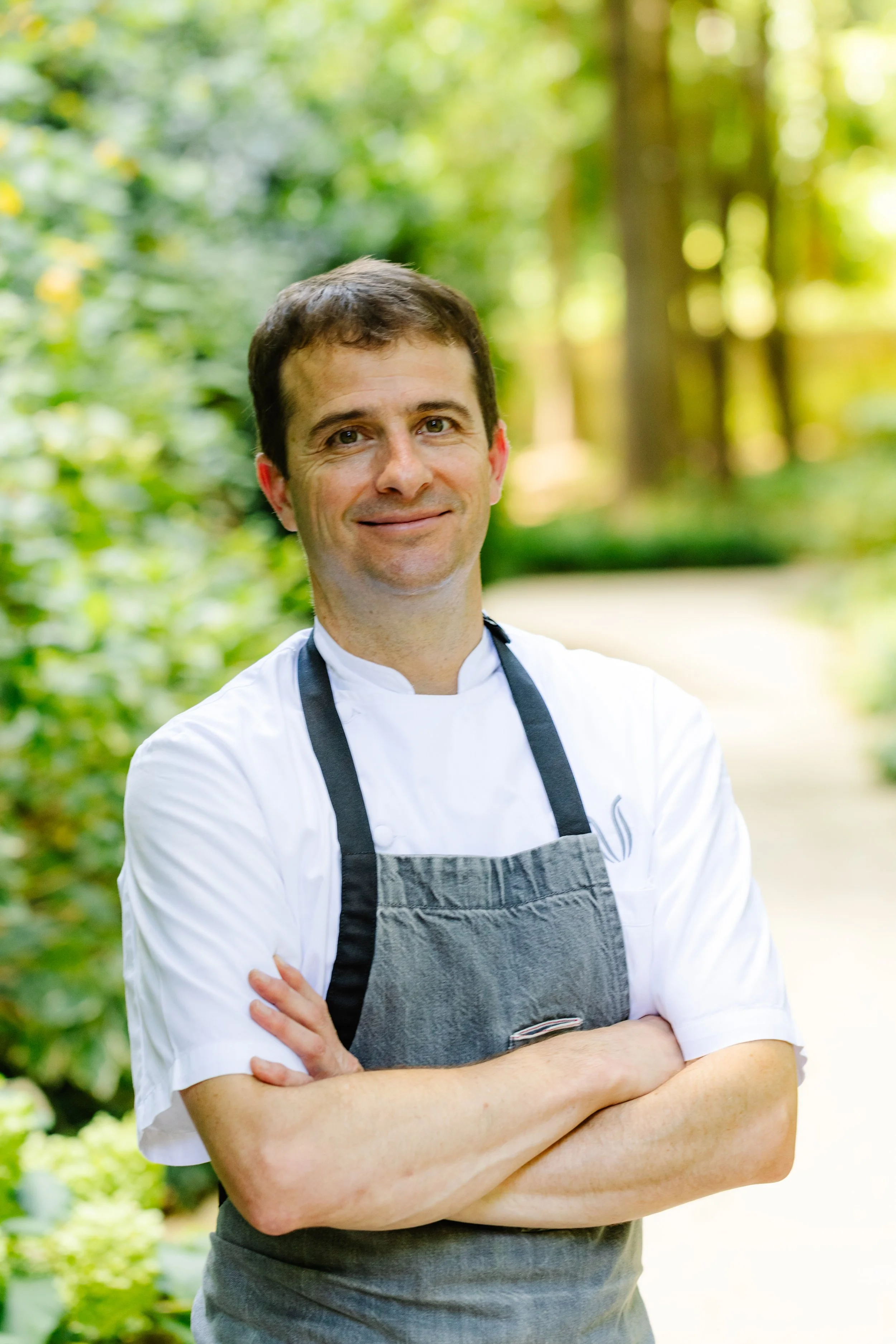 A male chef wearing a gray apron over a white chef's coat, standing outdoors with arms crossed and smiling, in a lush green park or garden.