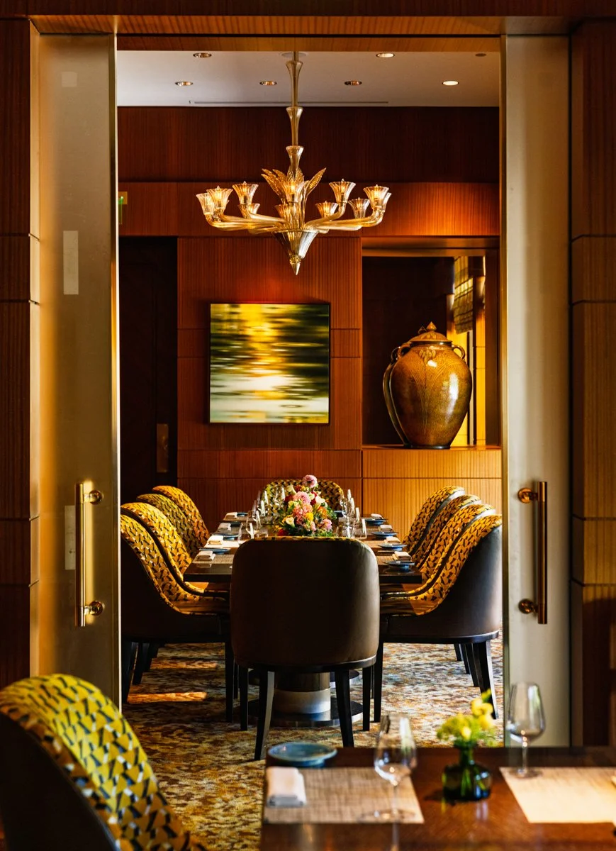 Elegant dining room with a long table set with floral centerpiece, surrounded by patterned chairs, a chandelier overhead, a large ceramic urn on a shelf, and artwork on the wall.