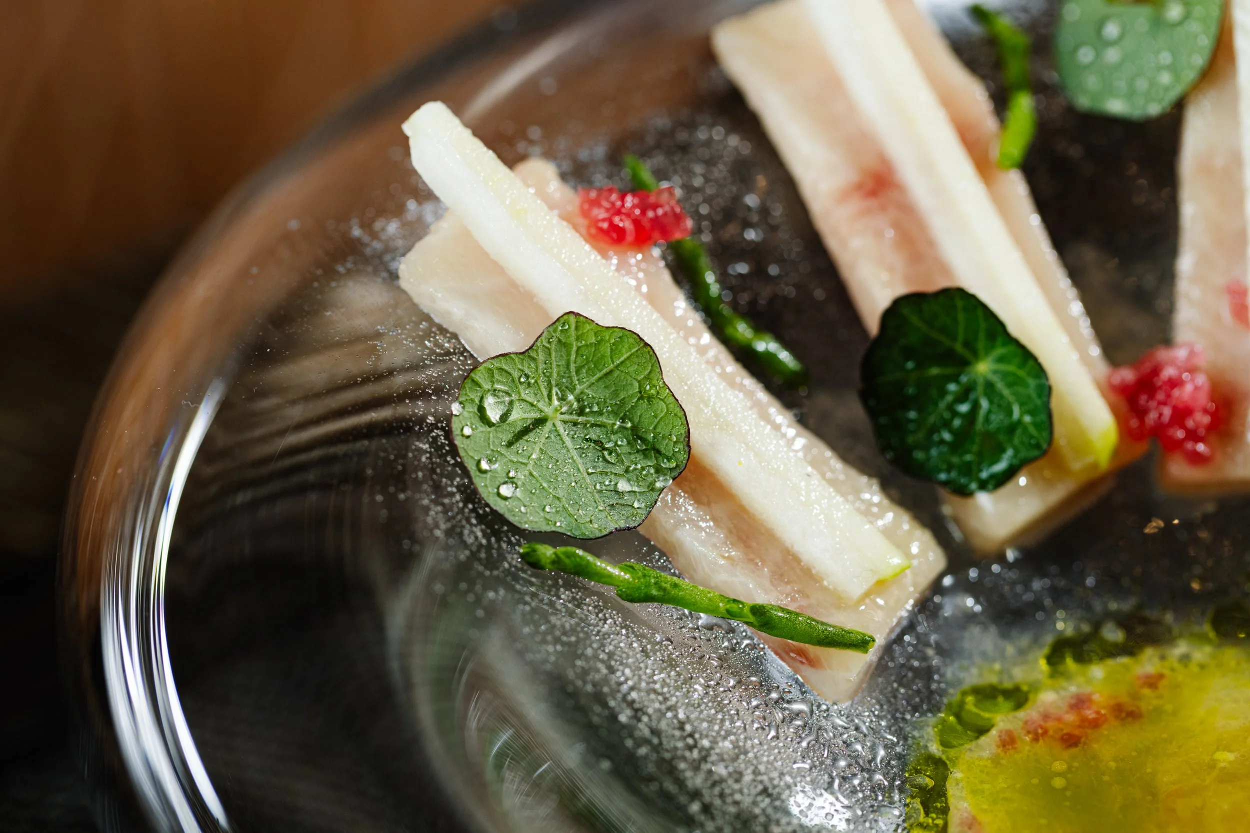 Close-up of a gourmet dish with thin slices of fish, green leaves, and small red garnishes on a dark plate, garnished with a green herb and a drizzle of sauce.