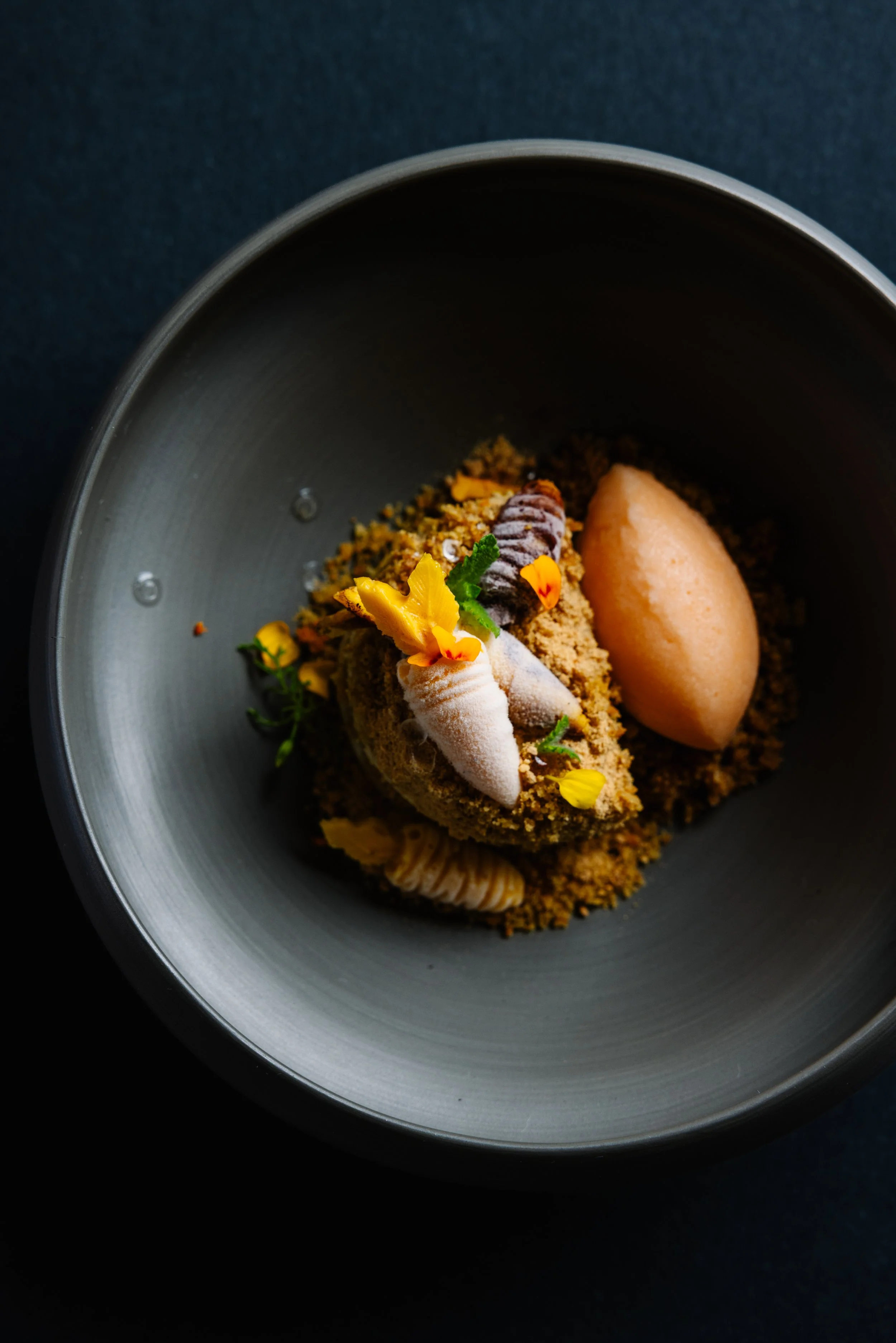 A plated dessert with a quenelle of light-colored sorbet, garnished with edible flowers and green herbs, resting on crumbled crumbly base with piped cream or mousse around the base.