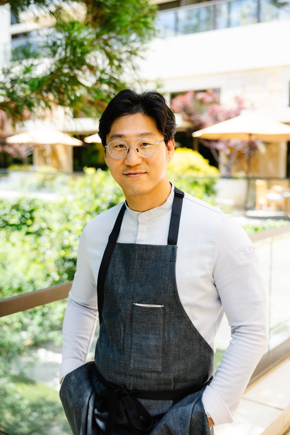 A man wearing glasses, a white shirt, and a dark apron, standing outdoors on a sunny day with greenery and umbrellas in the background.