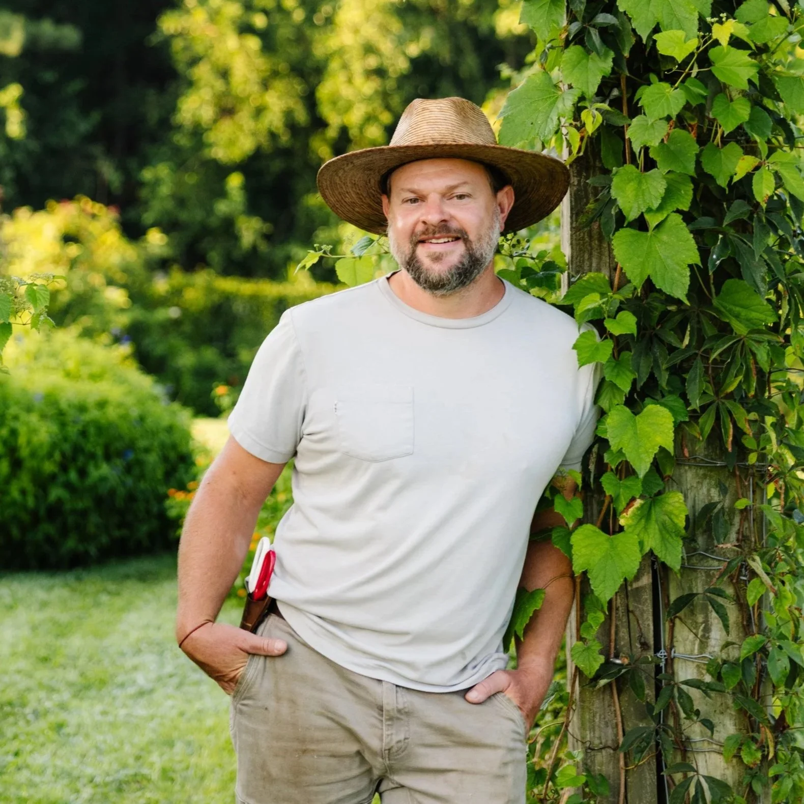 A man with a beard and mustache wearing a wide-brimmed straw hat and a light gray t-shirt, standing outdoors next to a vines-covered wooden post, smiling at the camera.
