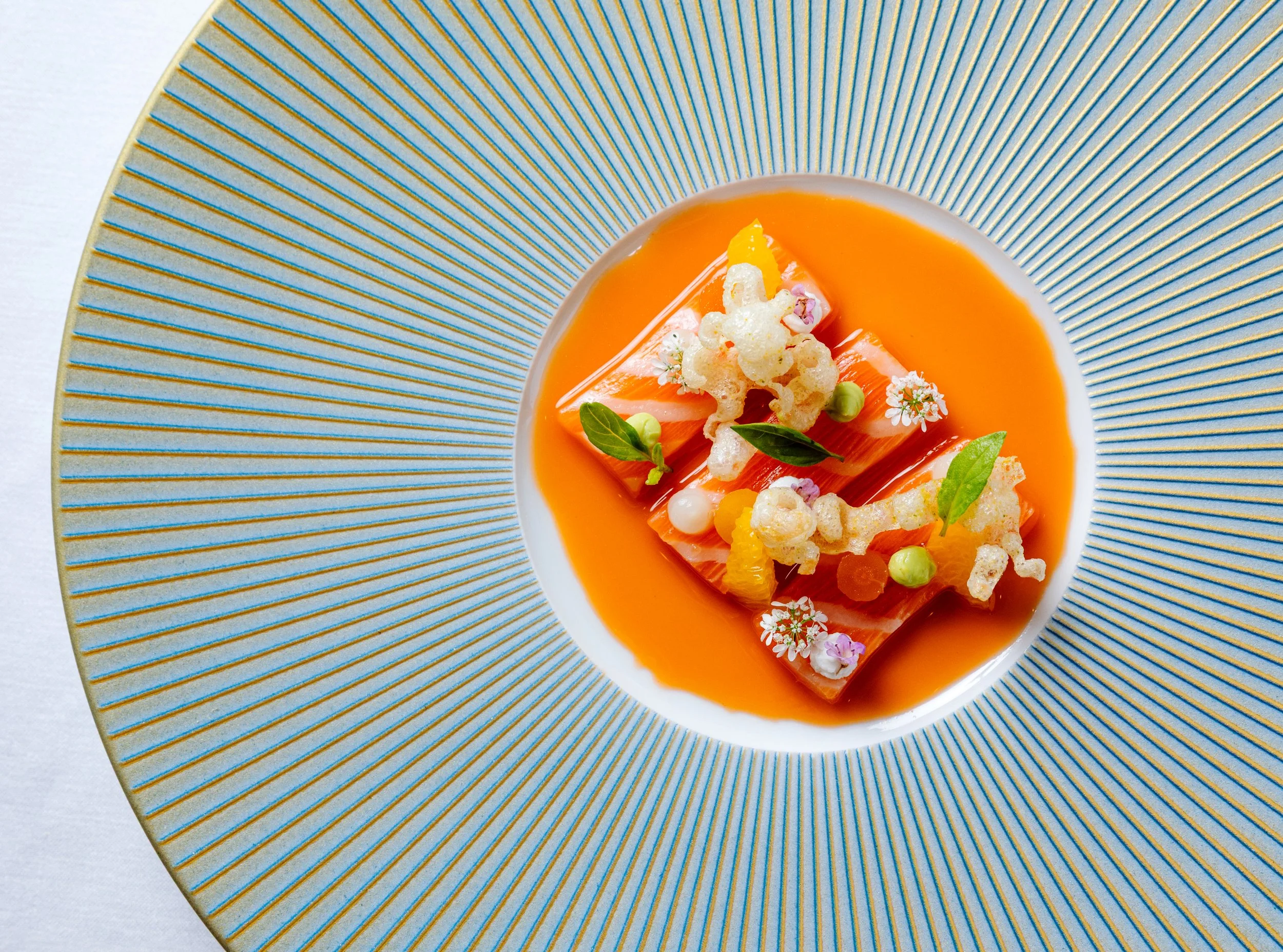 Elegant plated dish with thin slices of fish, orange sauce, edible flowers, microgreens, and crunchy garnishes on a decorative blue-rimmed plate.