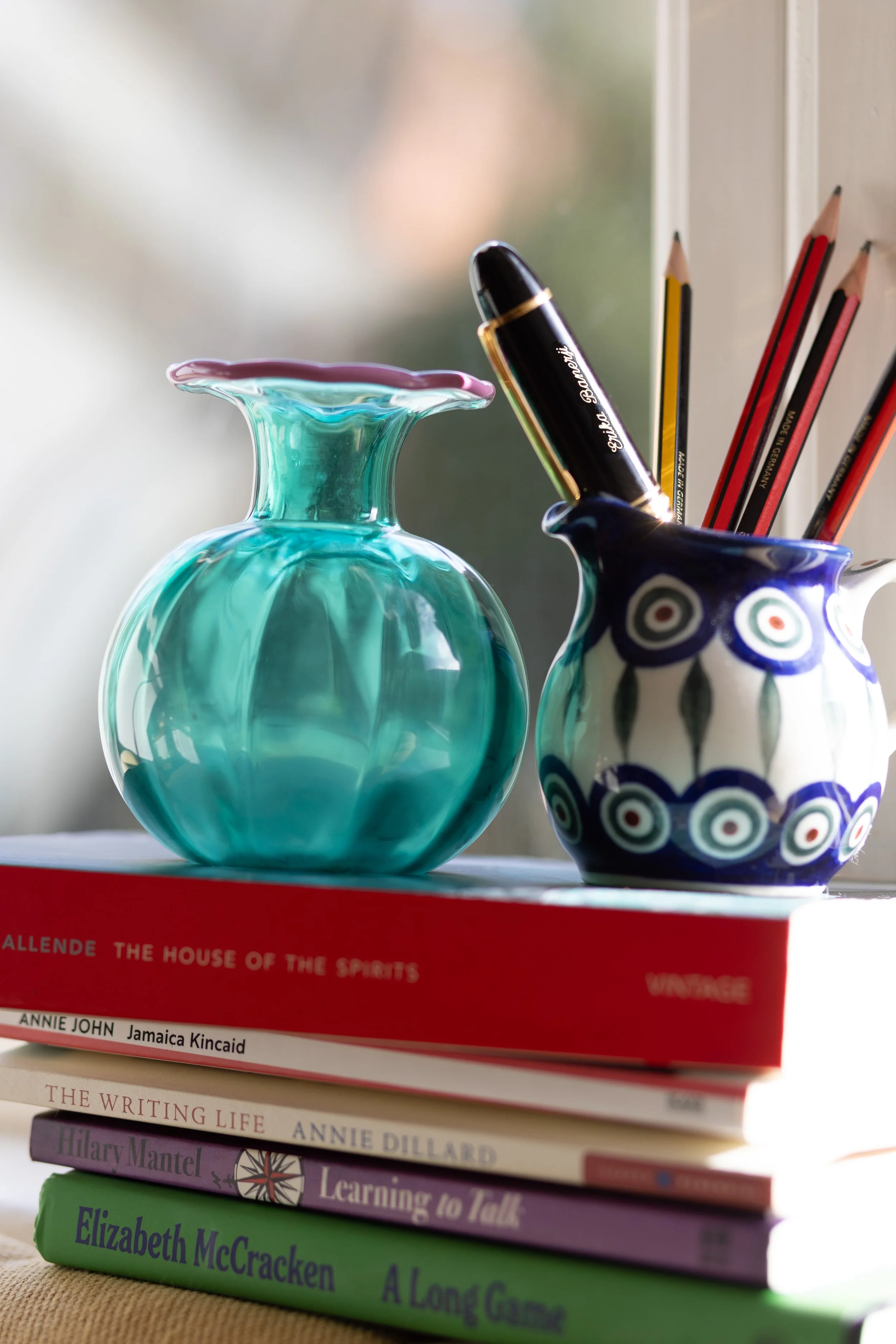 Stack of books with colorful ceramic vases and pens on top, placed on a table near a window.