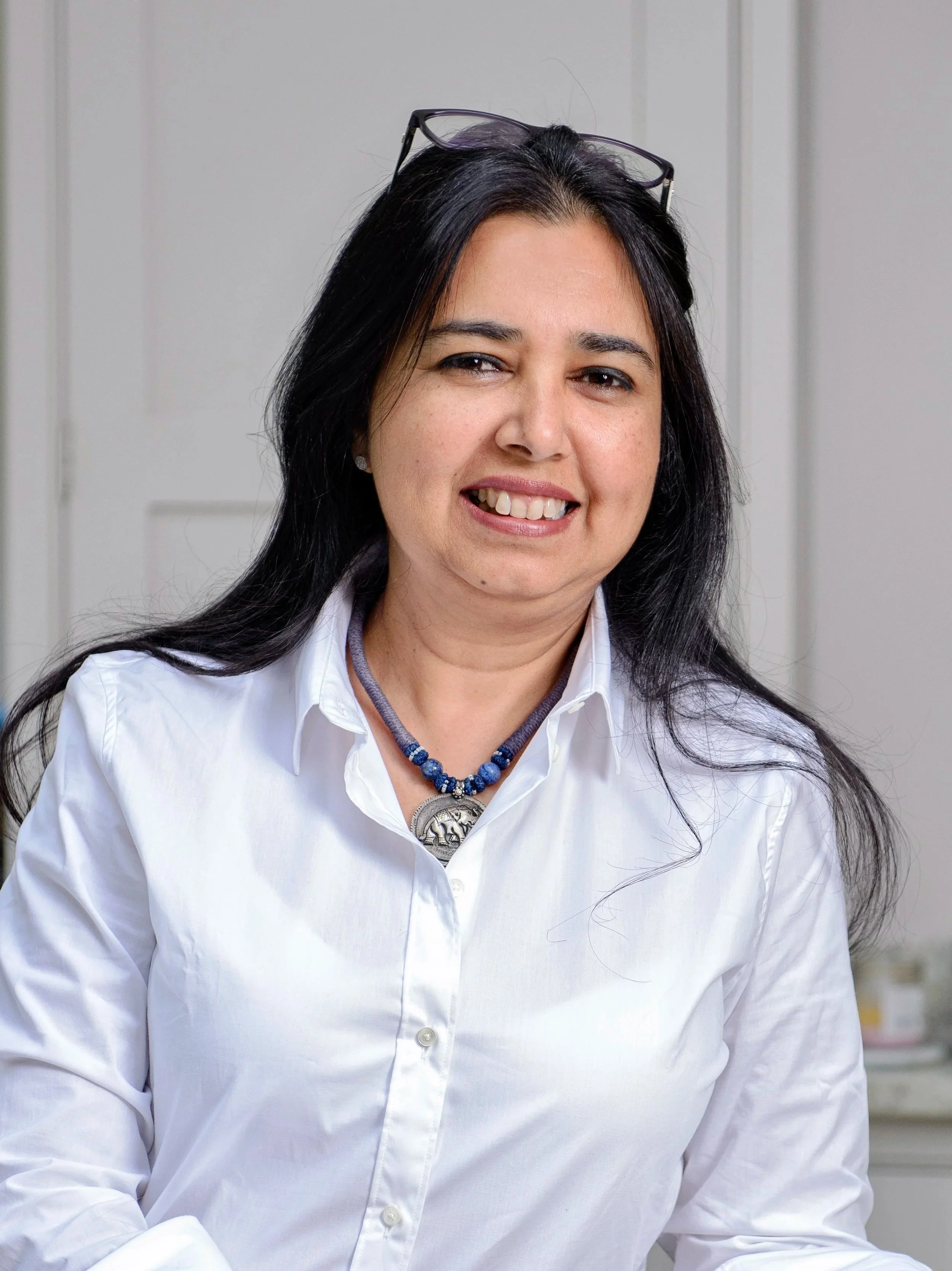 A woman with long black hair, wearing glasses on her head and a white button-up shirt, smiling and looking slightly to the right.
