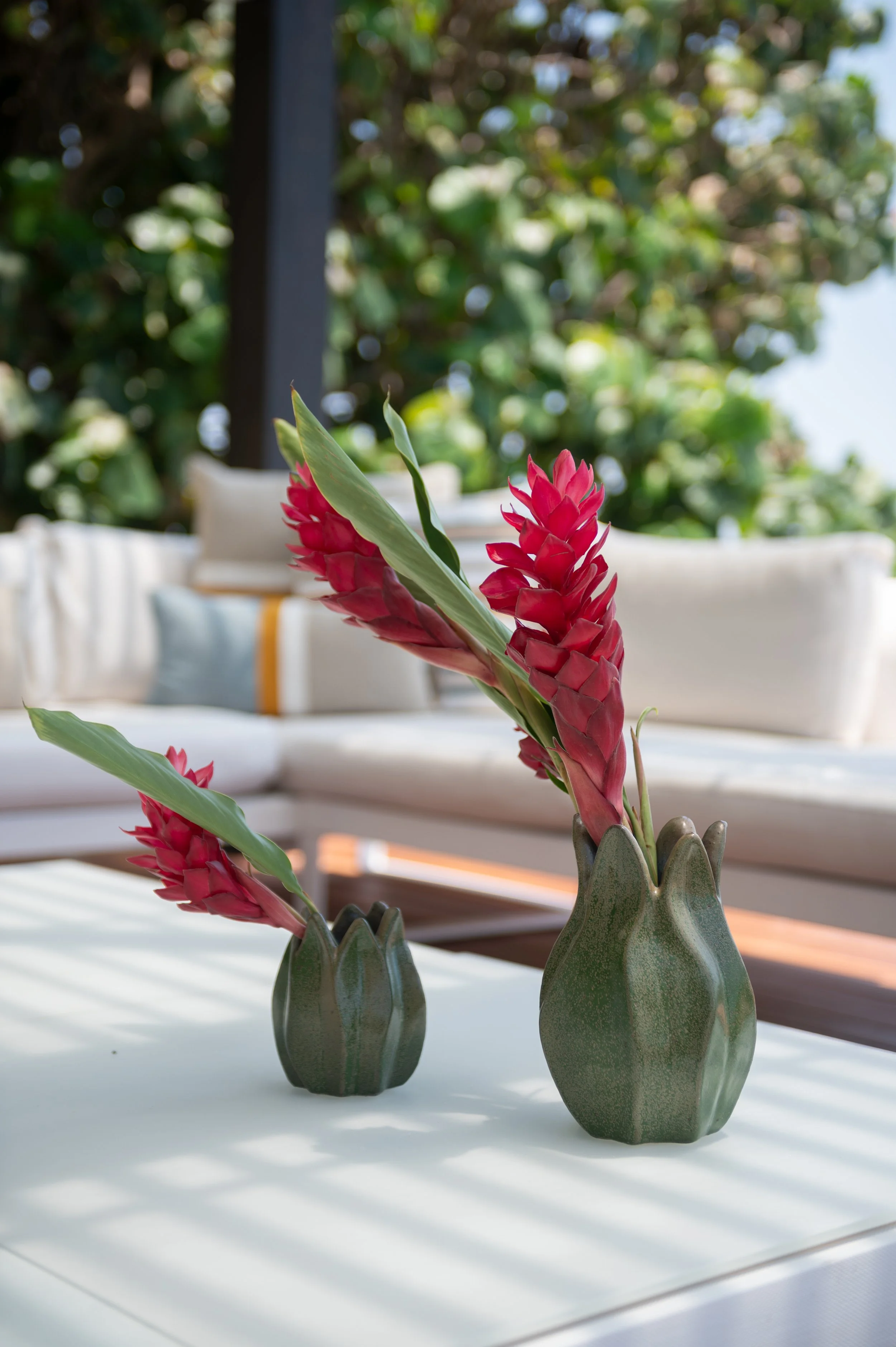 Two small green vases with pink tropical flowers on a white table, with blurred outdoor greenery in the background.