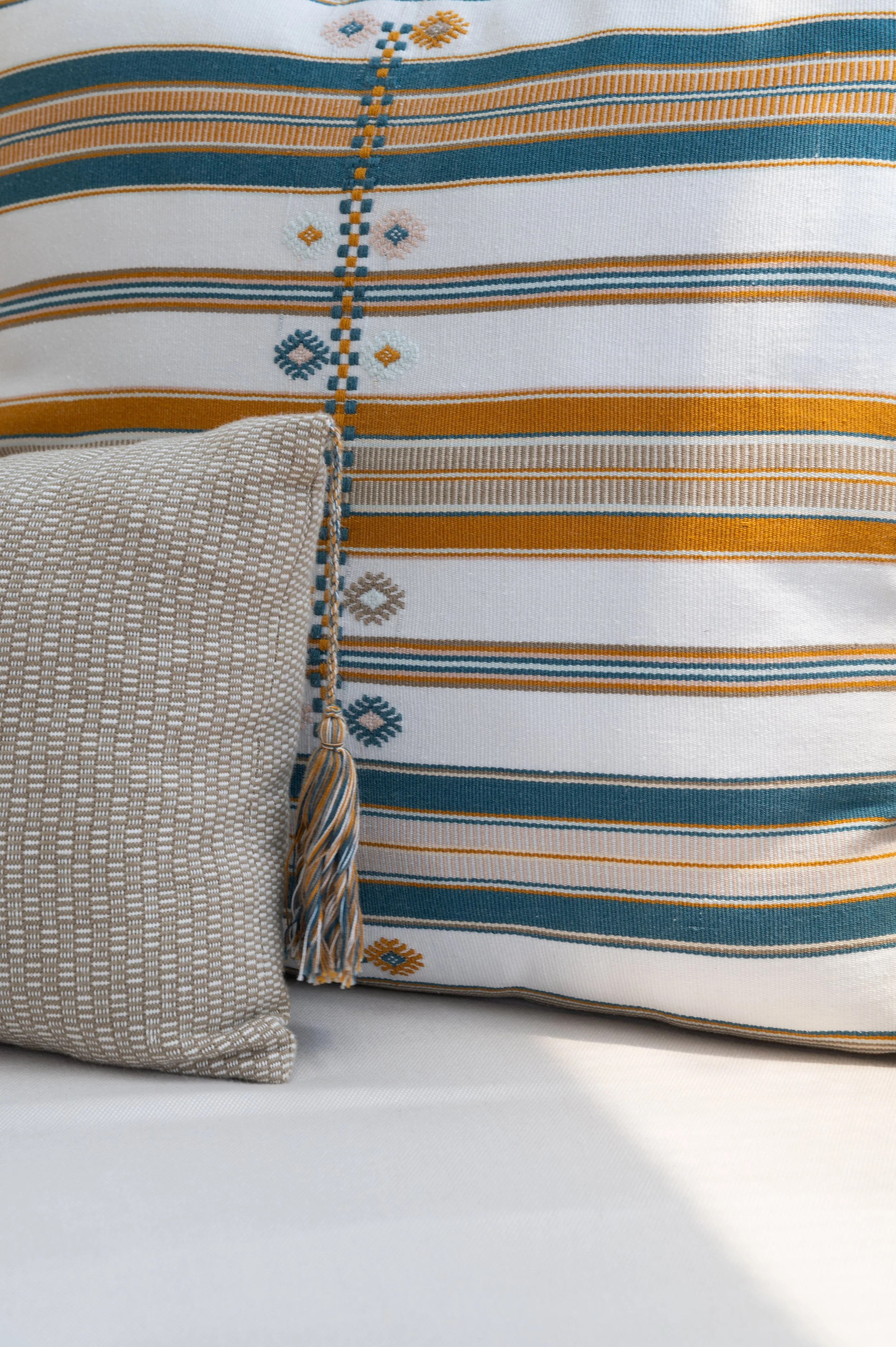 Close-up of two decorative pillows with striped and geometric patterns, one beige with a textured checkered design and the other white with orange, blue, and beige stripes, against a plain background.