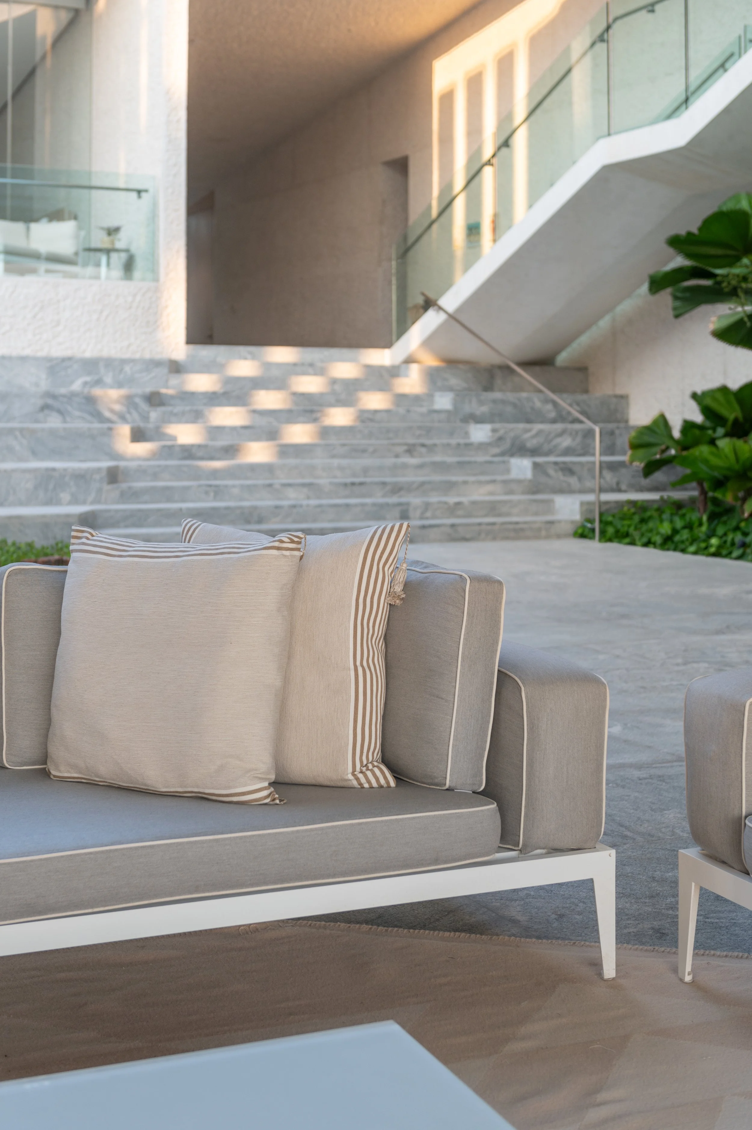 A modern outdoor seating area with beige cushioned furniture, striped pillows, concrete stairs, glass railing, green plants, and a staircase leading to an upper level with a white wall.