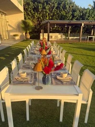 Long outdoor dining table set with white chairs, colorful floral arrangements, and place settings, in a grassy yard with trees and a covered patio in the background.