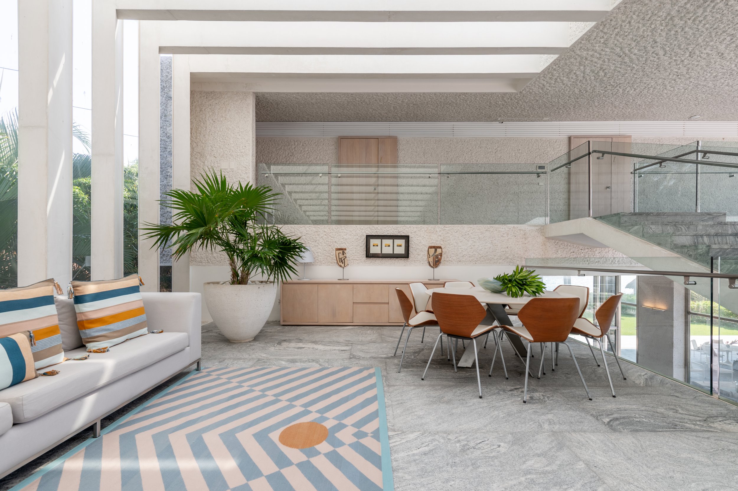 Modern living room with white sofa, colorful striped pillows, a geometric patterned rug, a large potted plant, and a dining table with wooden chairs, with a staircase and glass railing in the background.