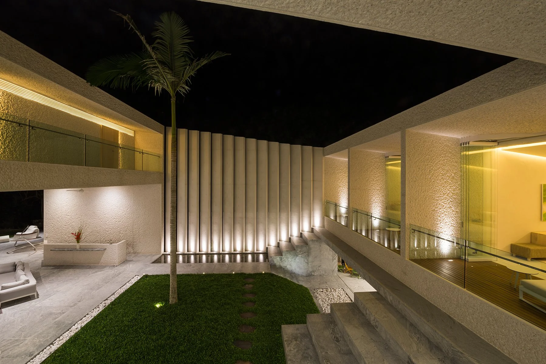 Modern two-story residence at night with illuminated exterior, glass railings, textured walls, and an outdoor seating area with a lounge chair, a small red flower arrangement, a manicured lawn, and a palm tree in the courtyard.