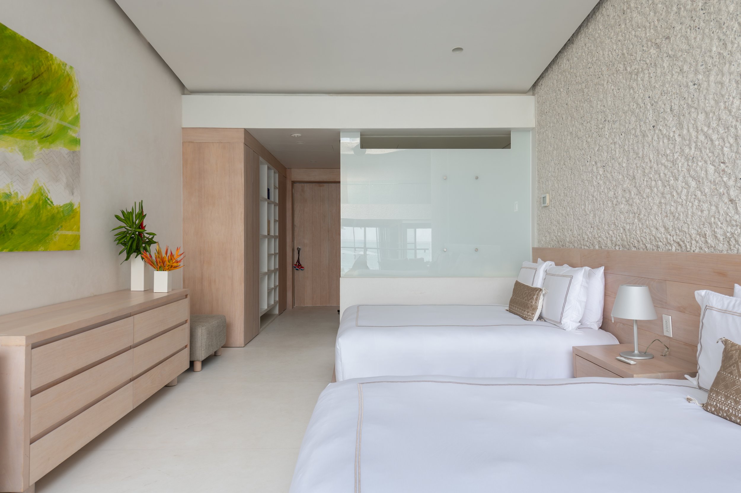 Modern bedroom with white walls, a wooden dresser, a bed with white linens, a bedside lamp, abstract green artwork, and potted plants.